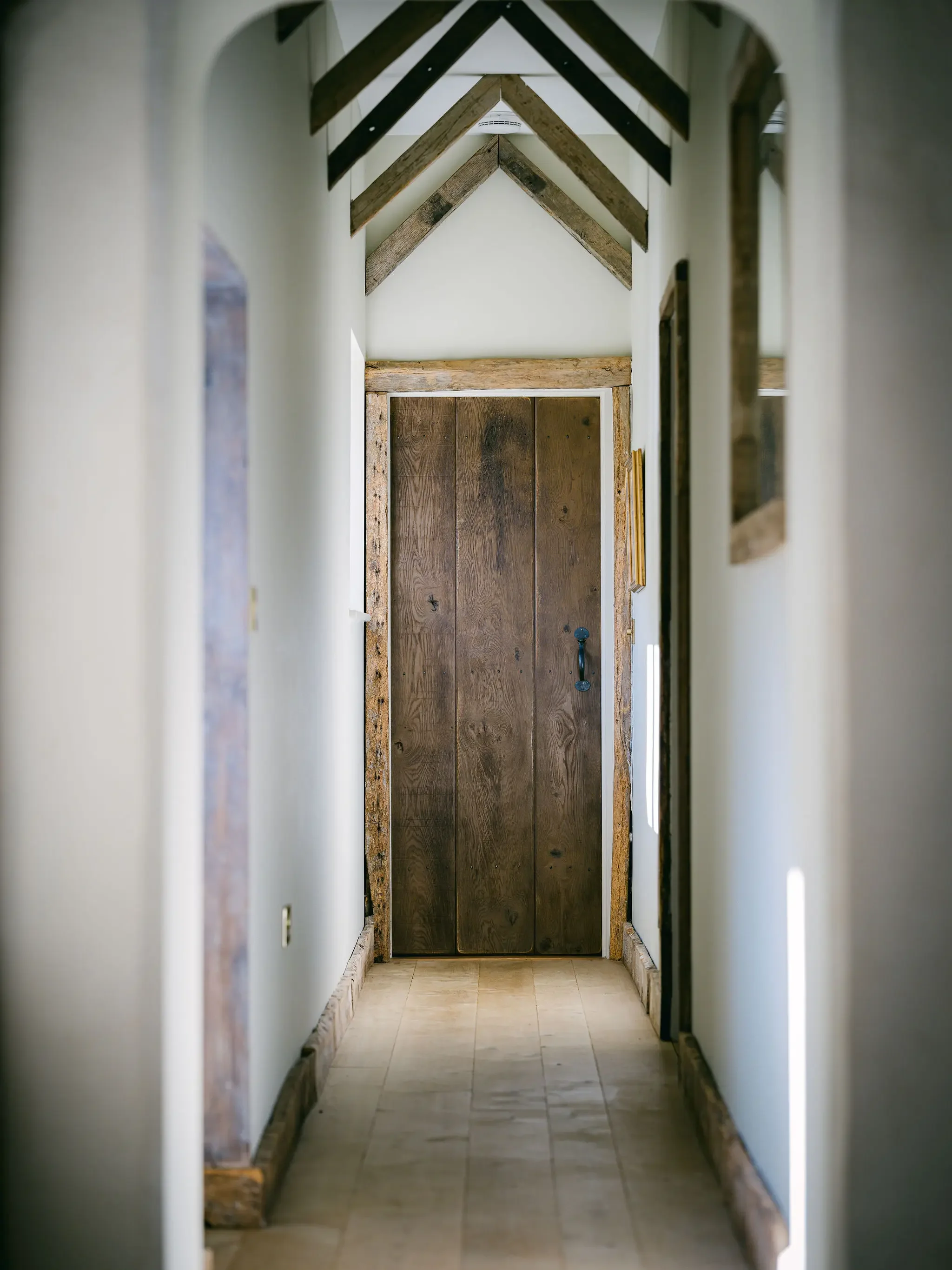 A narrow hallway with a wooden door at the end, white walls, and exposed wooden beams on the ceiling.