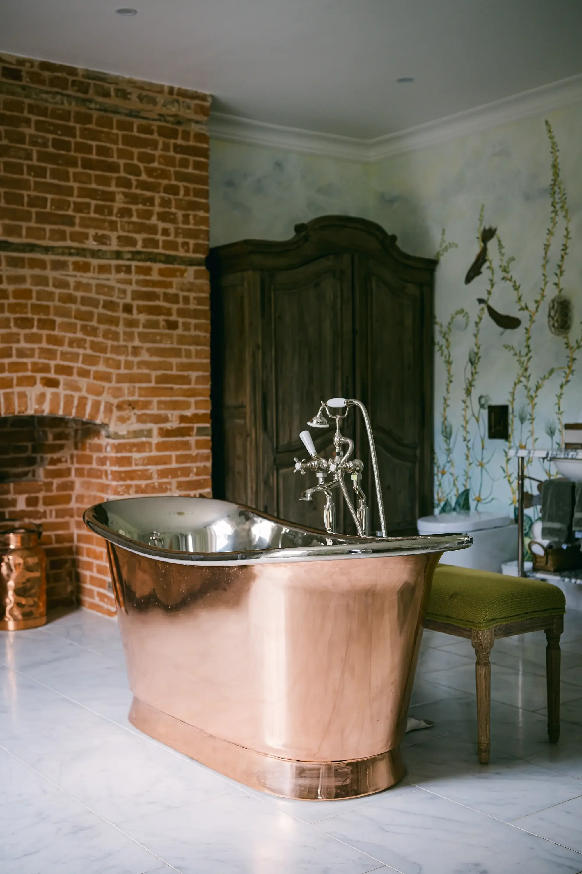 A bathroom with a copper bathtub, a brick wall on the left, a wooden armoire in the background, a mural of birds and plants on the wall, a green upholstered stool, and white tiled flooring.