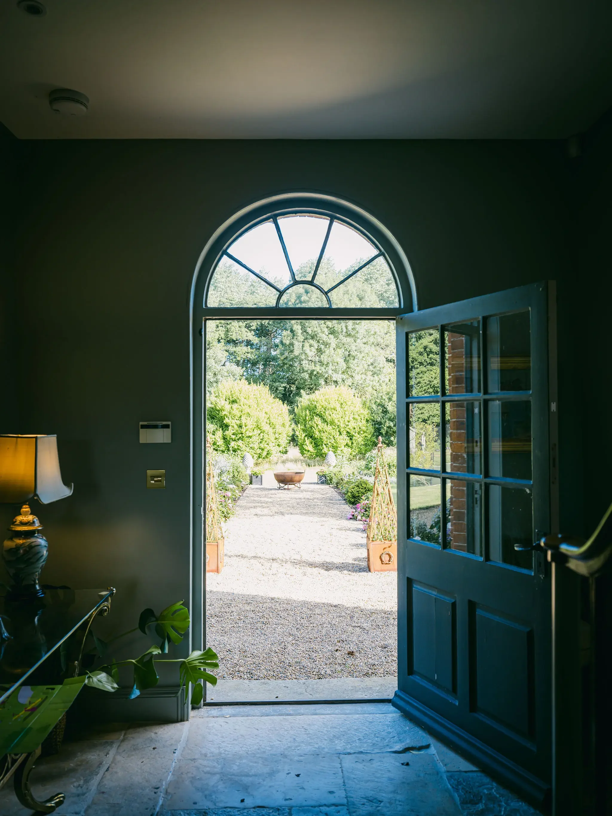 View through an open door showing a garden with trees, potted plants, and a gravel pathway outside, with a clear sky in the background.