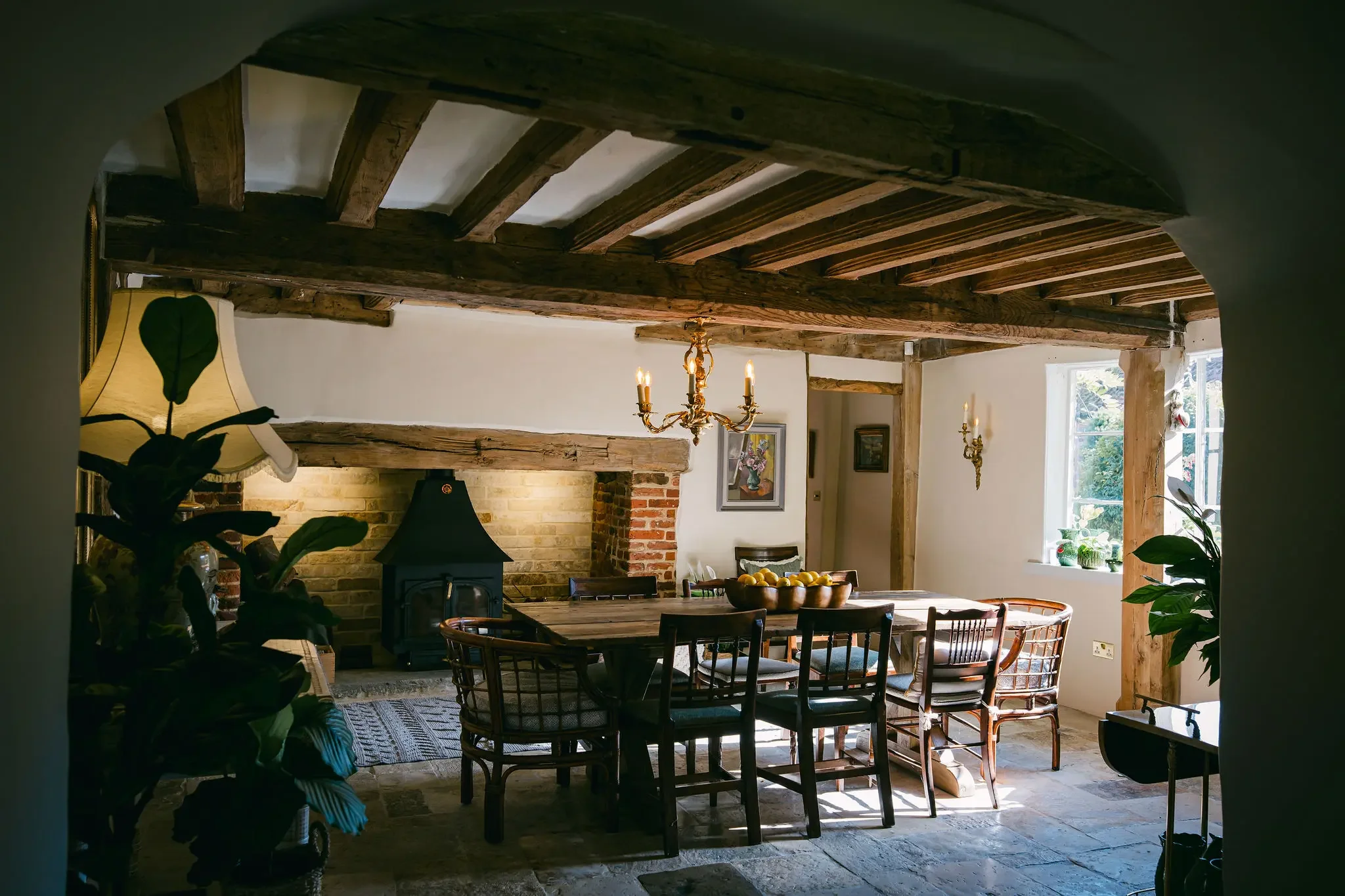 A cozy dining room with a rustic design, featuring wooden ceiling beams, a chandelier, and a brick fireplace. There is a wooden dining table with chairs, a large bowl of lemons, and a window with sunlight streaming in, illuminating potted plants and wall art.