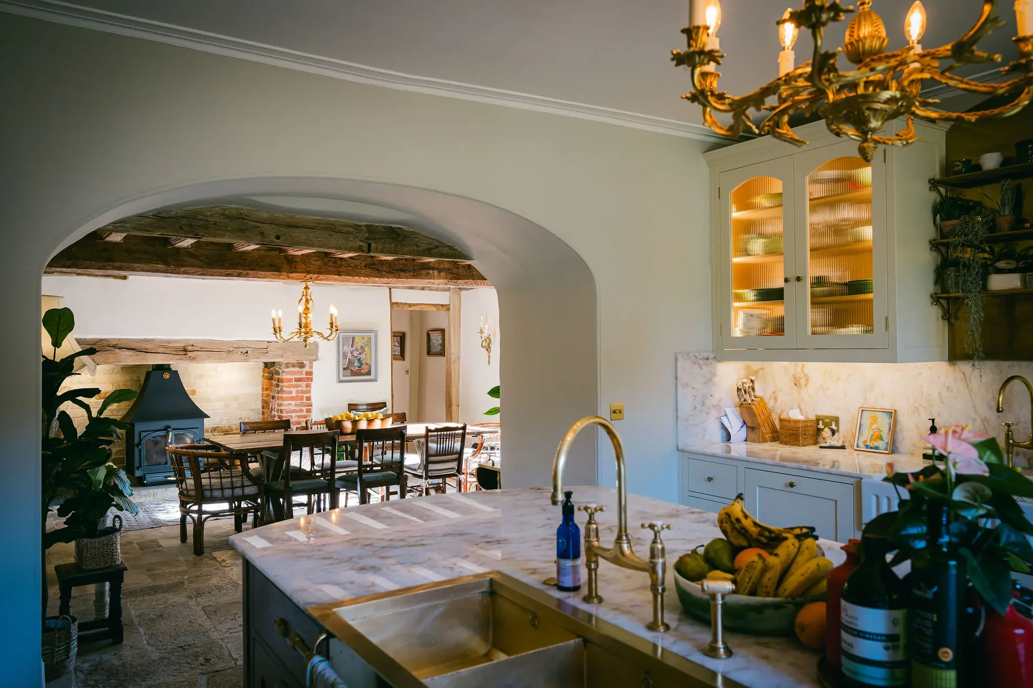 Kitchen with a white marble island, a sink with a gold faucet, and a bowl of bananas and other fruits. Open-concept dining and living area with a brick chimney, wooden beams, and a chandelier. Green houseplants and various decorations visible.