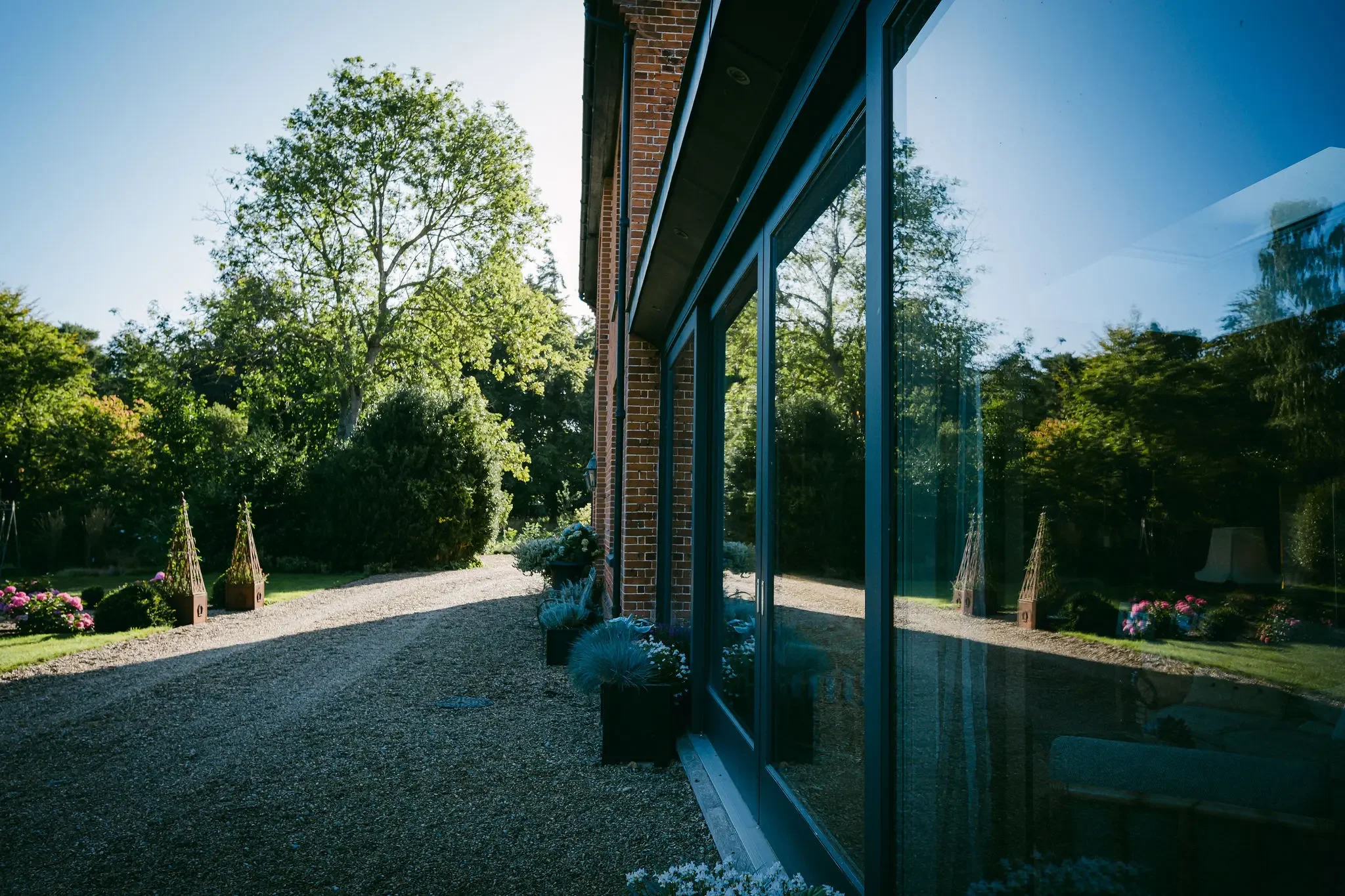 Sunlit garden with trees and flowering plants viewed from beside a brick building with large glass windows.