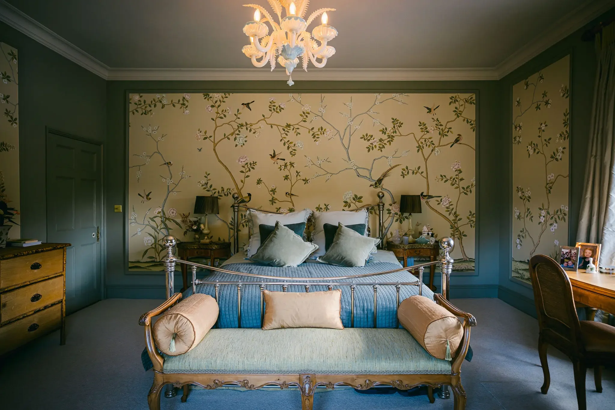 A bedroom with a metal bed featuring pillows and a bedspread, an antique wooden chaise lounge at the foot, a golden-topped bedside table with framed photos, black lamps on the nightstands, a large floral and bird mural on the wall behind the bed, a chandelier hanging from the ceiling, and a window with curtains on the right side.