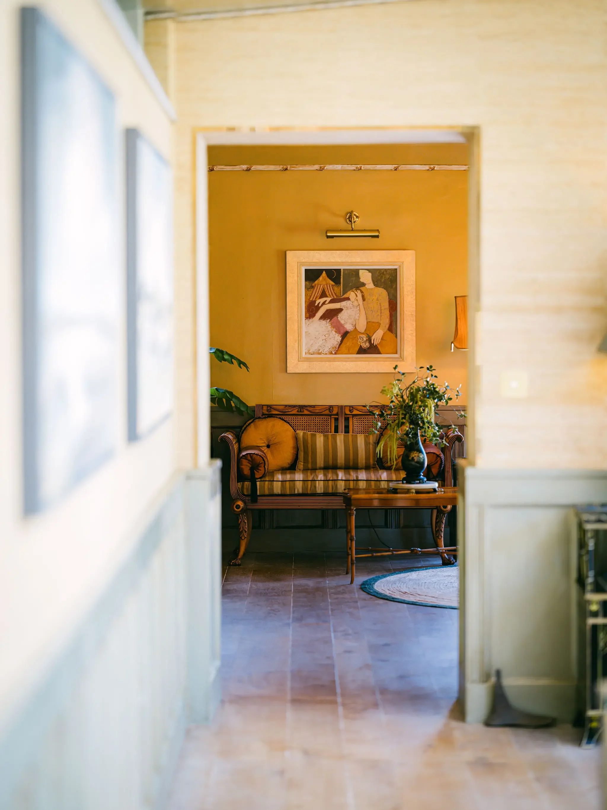 View through a doorway into a living room with a vintage-style couch, a gold-framed painting on the wall, a plant, a side table, and a small circular rug.