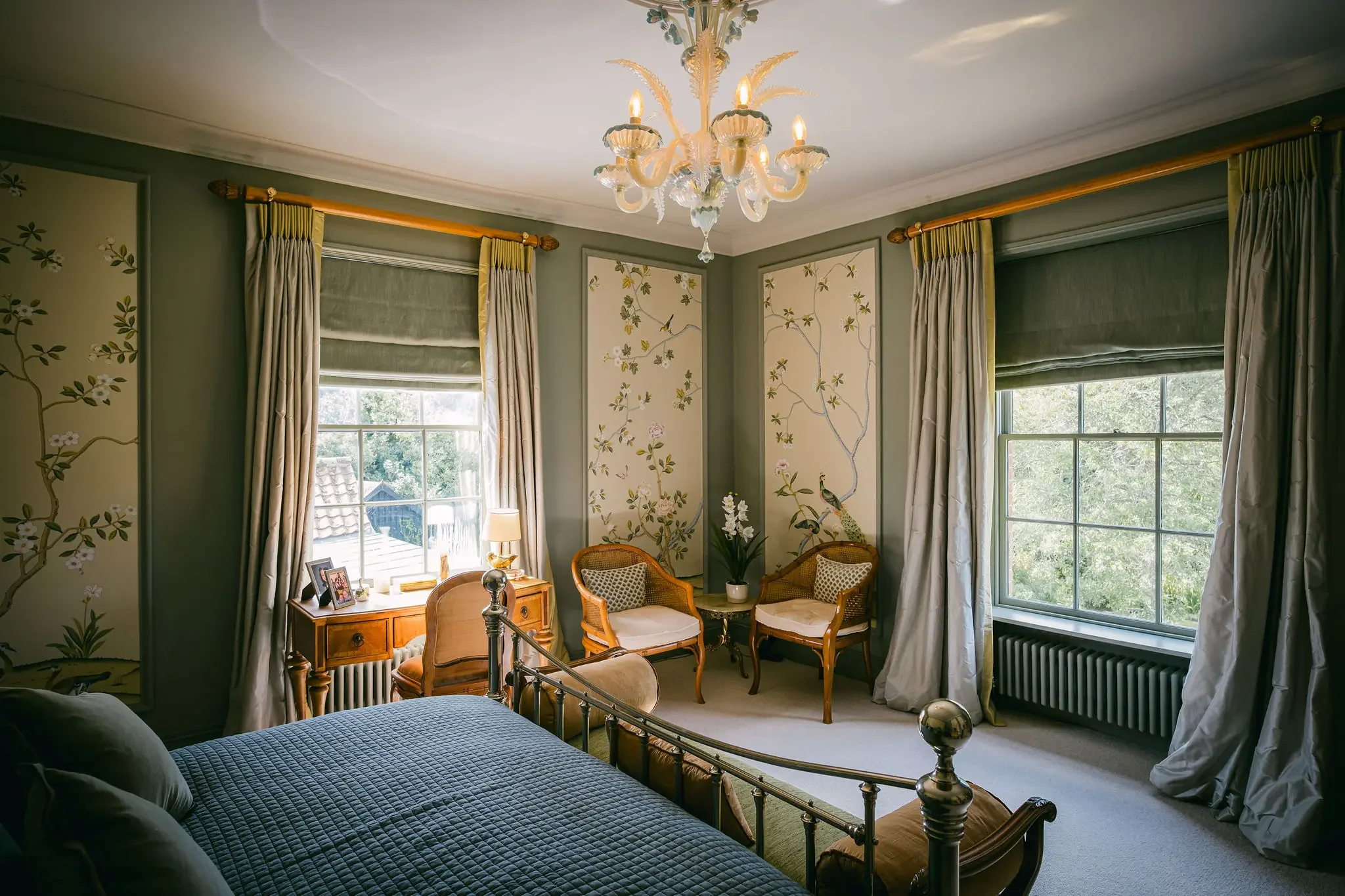 Elegant bedroom with a vintage brass bed, floral wallpaper, a wooden writing desk, and two armchairs near windows with cream and green curtains, and a chandelier hanging from the ceiling.