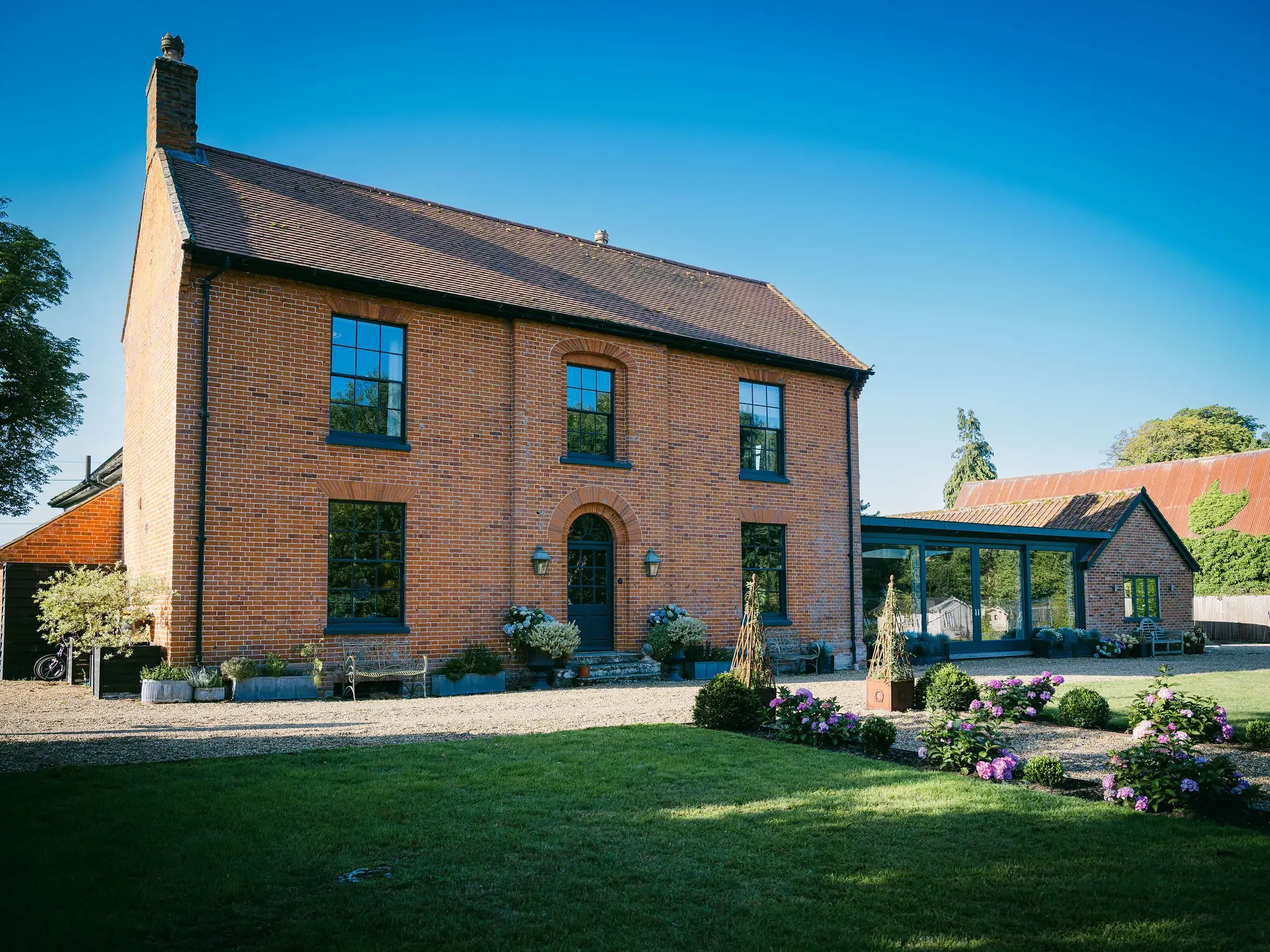A red-brick house with large windows and a sloped roof, surrounded by a garden with purple flowers, green bushes, and trees under a clear blue sky.