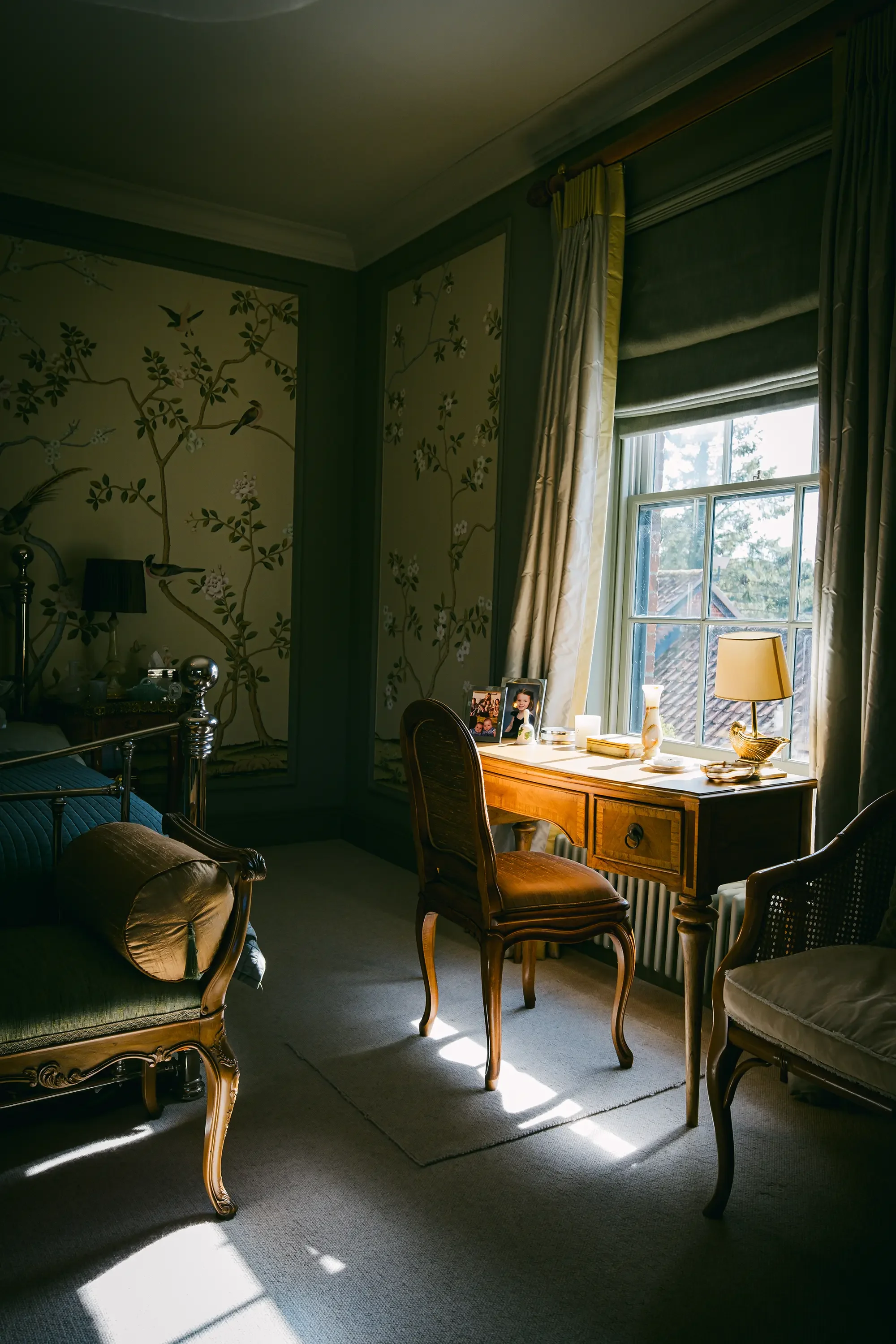 A cozy sitting room with antique furniture and a window letting in natural light. The room has floral wallpaper with birds, a wooden desk with framed photos, candles, and decorative items, along with a mix of upholstered chairs and a sofa. The curtains are pulled aside to reveal a view of trees and rooftops outside.