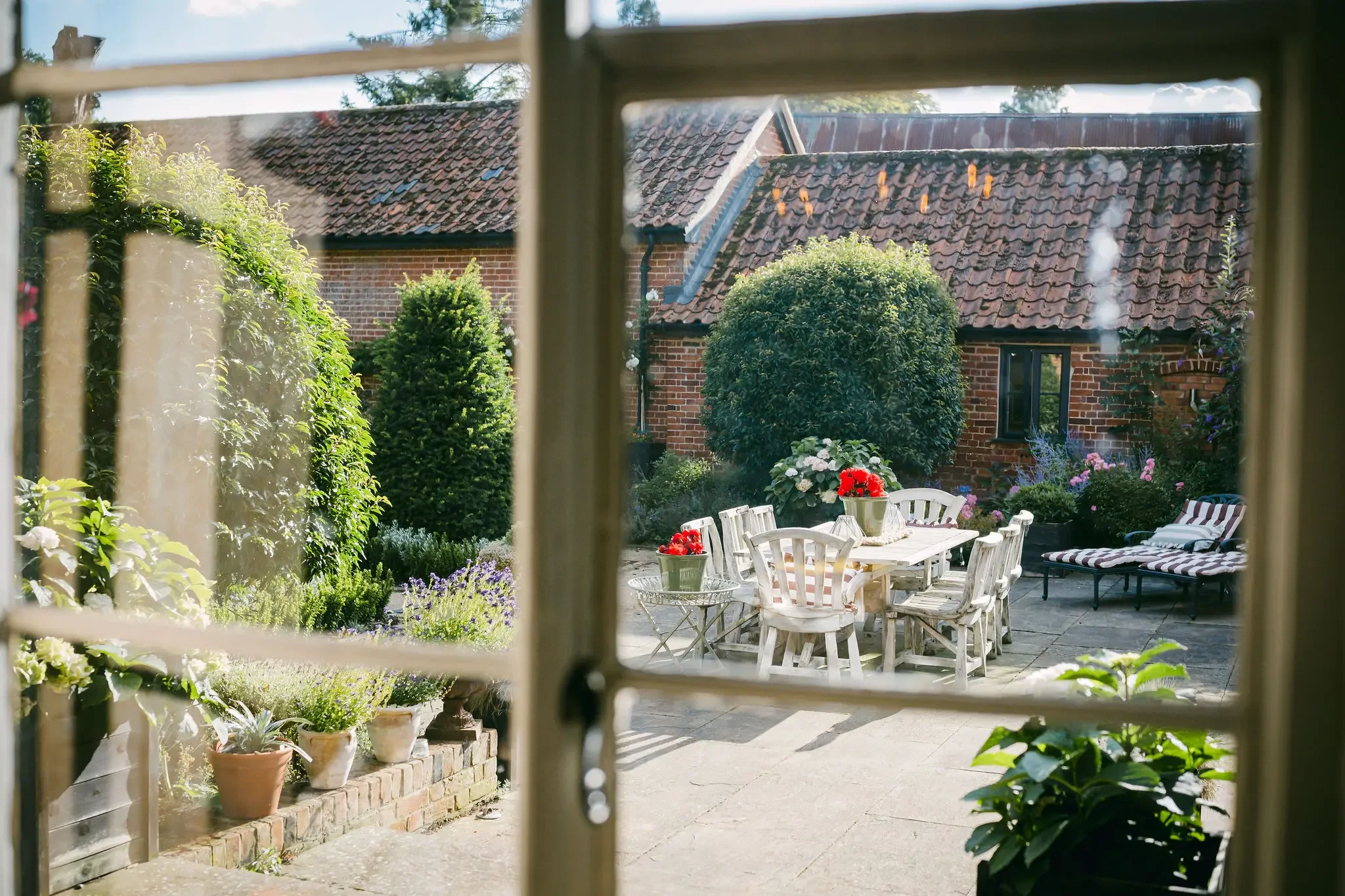 View of a backyard garden with outdoor furniture, potted plants, bushes, and a brick house with a tiled roof, seen through a partially open window.
