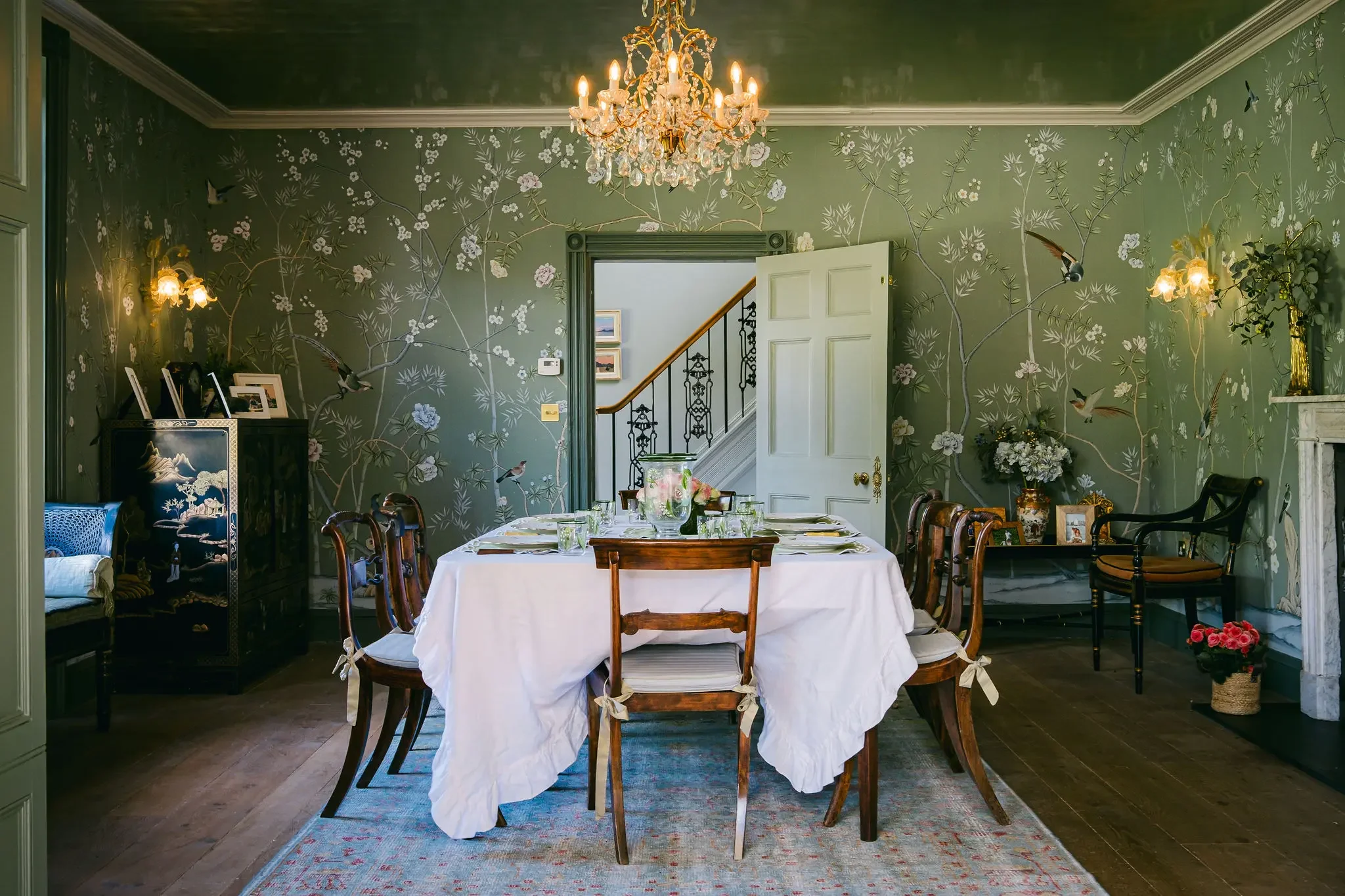 A dining room with a round table covered with a white tablecloth, set with glasses and dishes. The room has green floral wallpaper, a chandelier hanging from the ceiling, and wall-mounted lamps. There is a doorway leading to a staircase and other framed pictures on the wall.