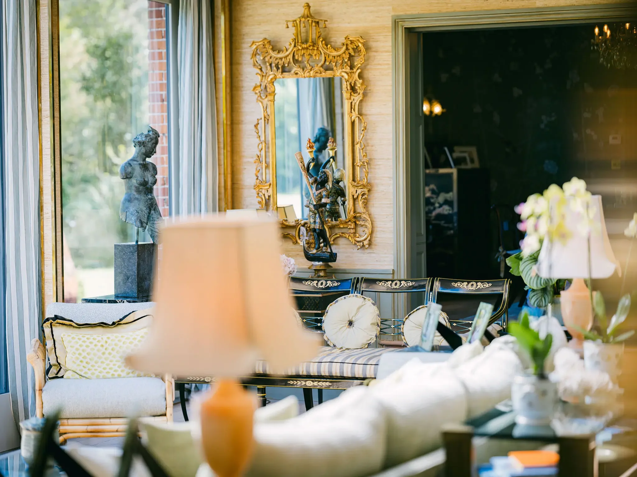 A warmly lit room with elegant decor, including a bronze bust sculpture, a large ornate gold mirror, and a display of decorative medieval knights, with windows and curtains in the background.