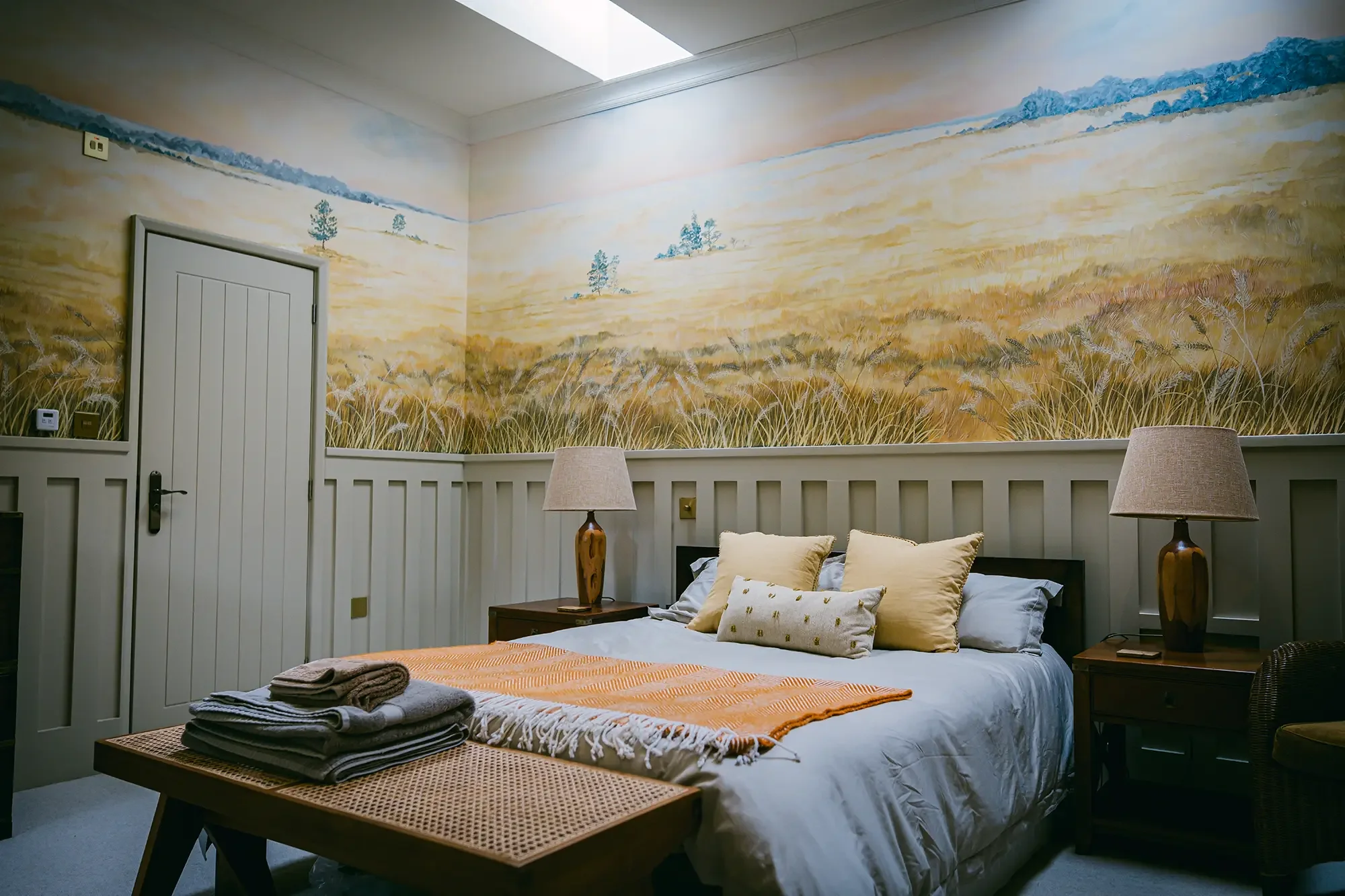 A cozy bedroom with a large scenic landscape mural on the wall above the bed, featuring rolling fields and distant mountains; the bed has multiple pillows and a throw blanket. Two bedside tables with lamps are beside the bed, and a wooden table at the foot of the bed holds folded towels.