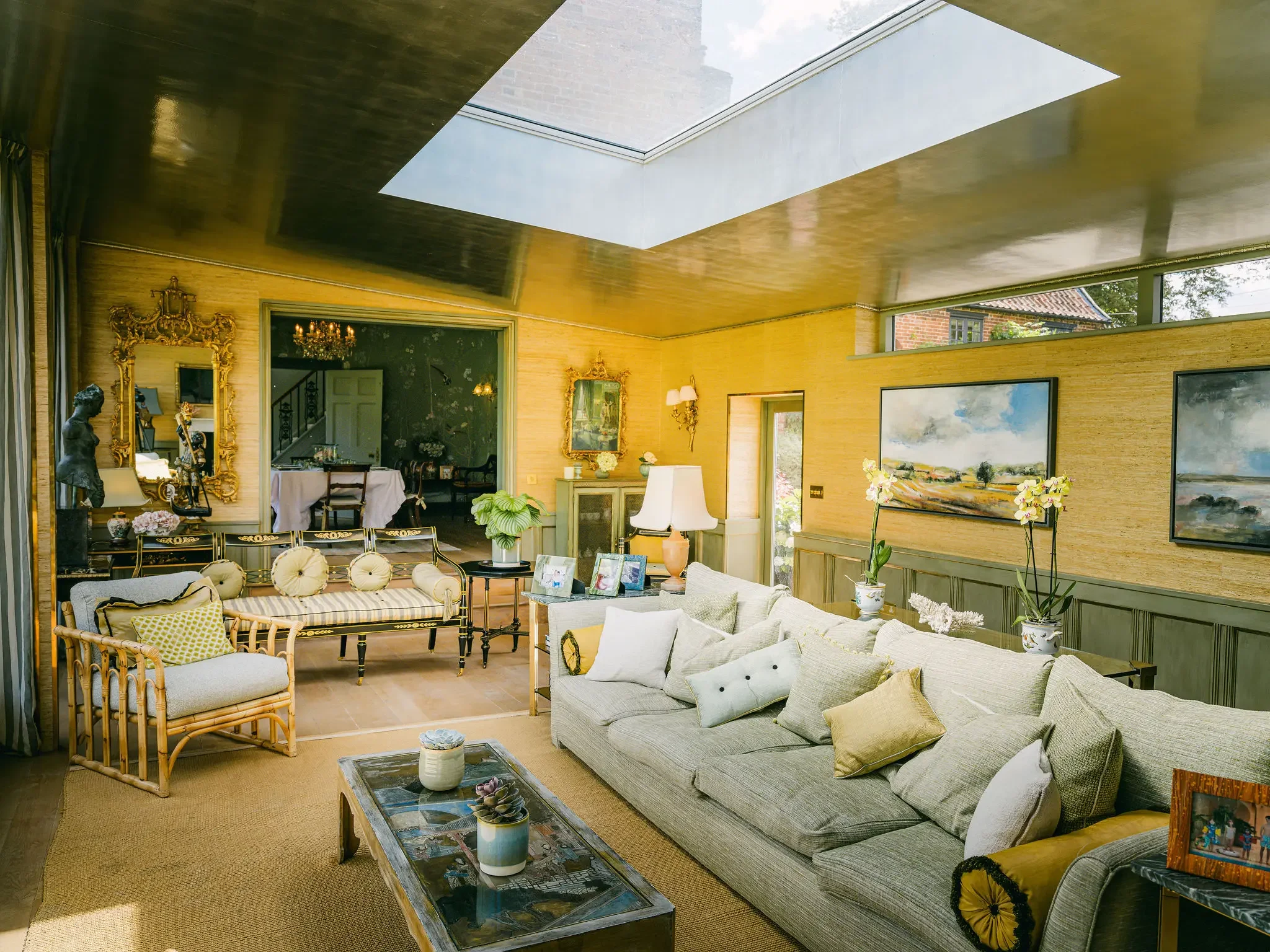 A spacious living room with beige walls, a large L-shaped light-colored sofa with multiple pillows, a rectangular coffee table with decorative items, and a separate sitting area with chairs. The room has large windows with views of trees and a neighboring building, a gold-framed mirror, a chandelier, and artwork on the walls. There are plants and decorative objects throughout the space, with sunlight coming through a large skylight in the ceiling.