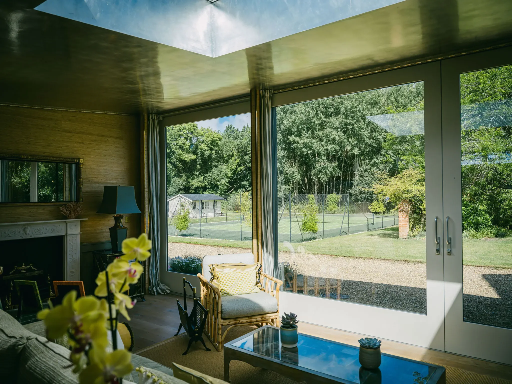 Interior of a living room with large glass doors opening to a backyard tennis court and lush green trees, furnished with a wicker armchair with cushion, a coffee table with potted plants, and framed photographs on a table.