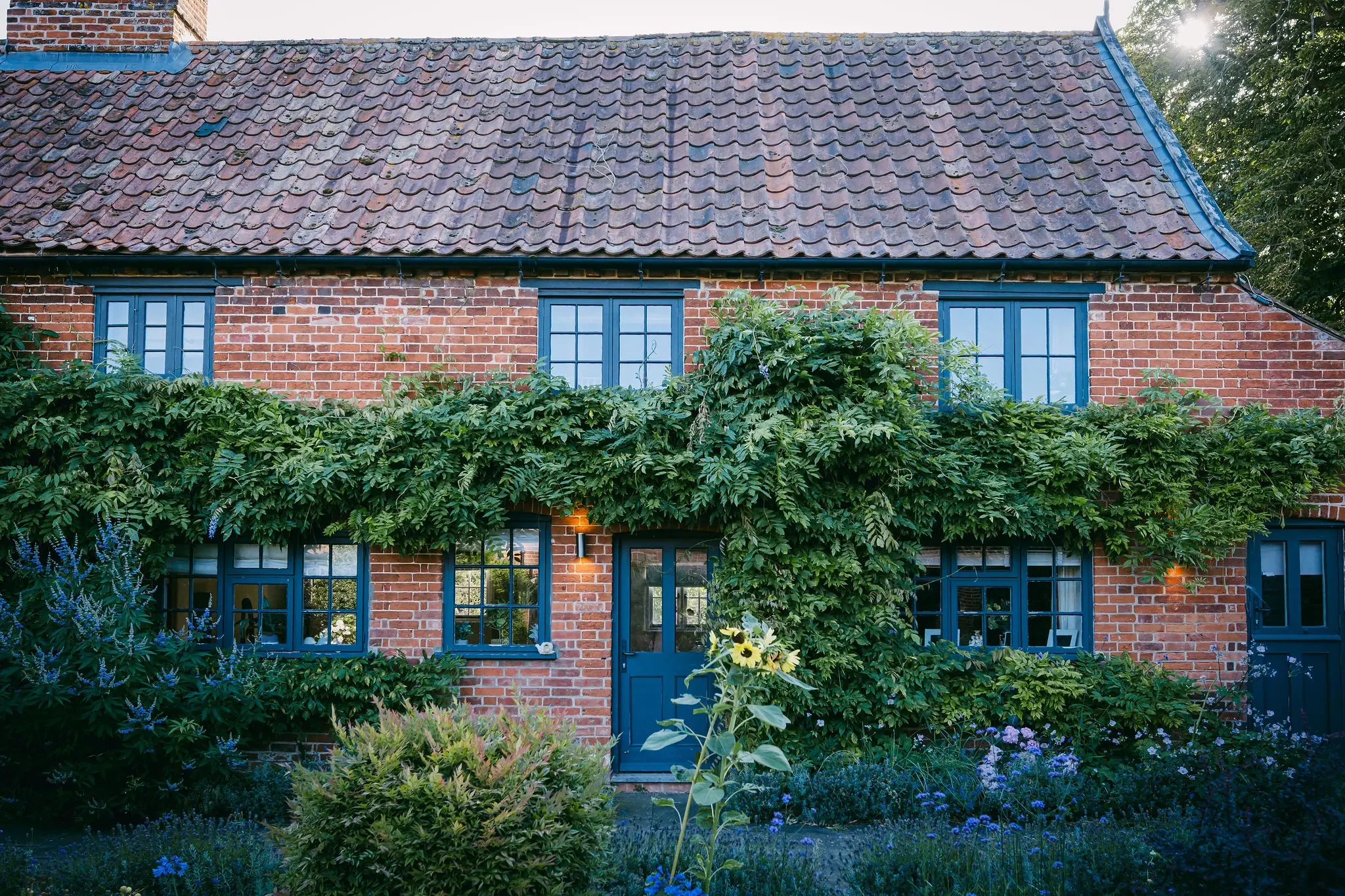 A brick house with a tiled roof and blue window frames, surrounded by a lush garden with various plants and flowers.