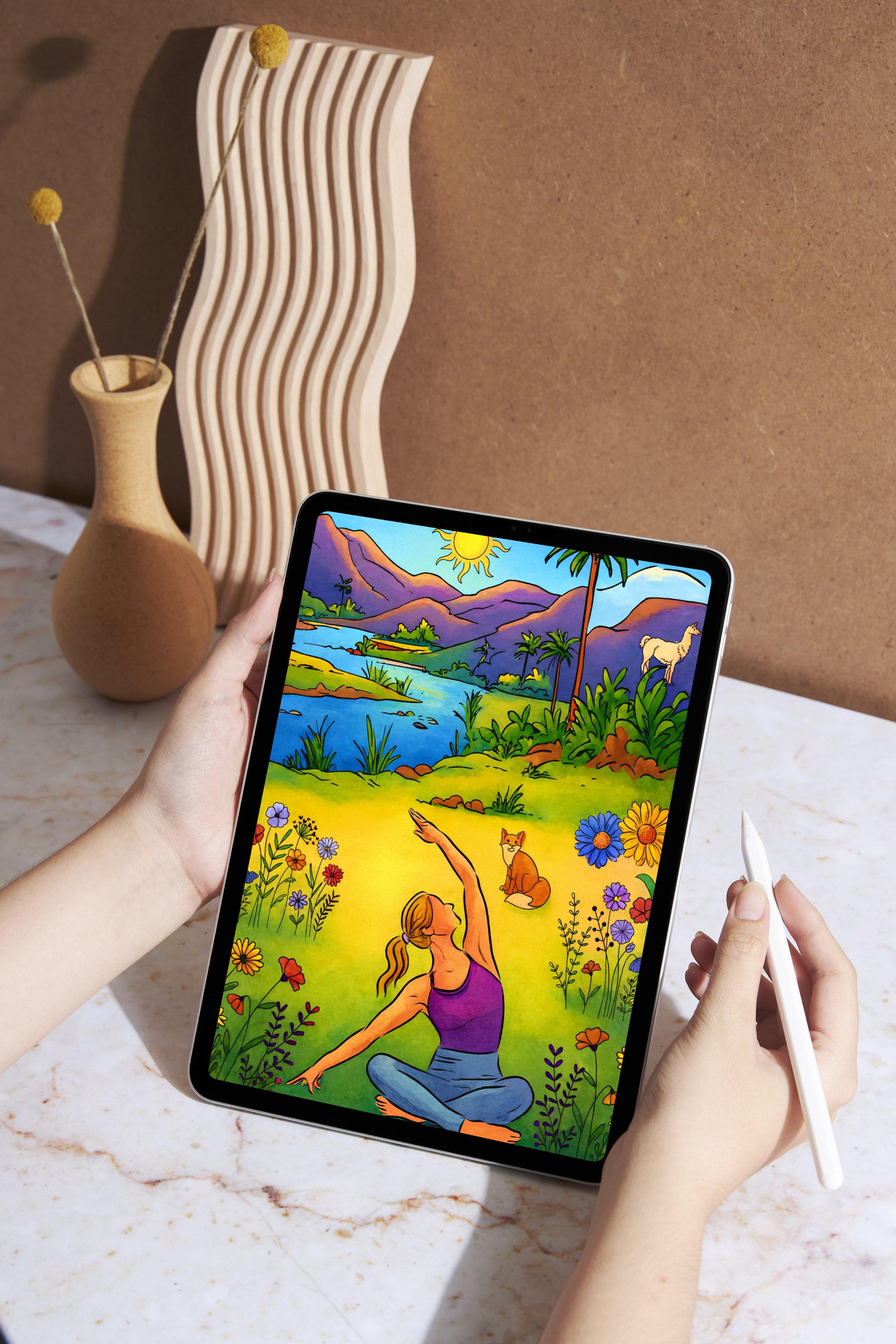 Person holding a tablet with an illustration of a woman practicing yoga outdoors amidst colorful flowers, with a mountain lake and green landscape in the background. A stylized vase with dried flowers and a decorative wooden sculpture are in the background on a marble surface.
