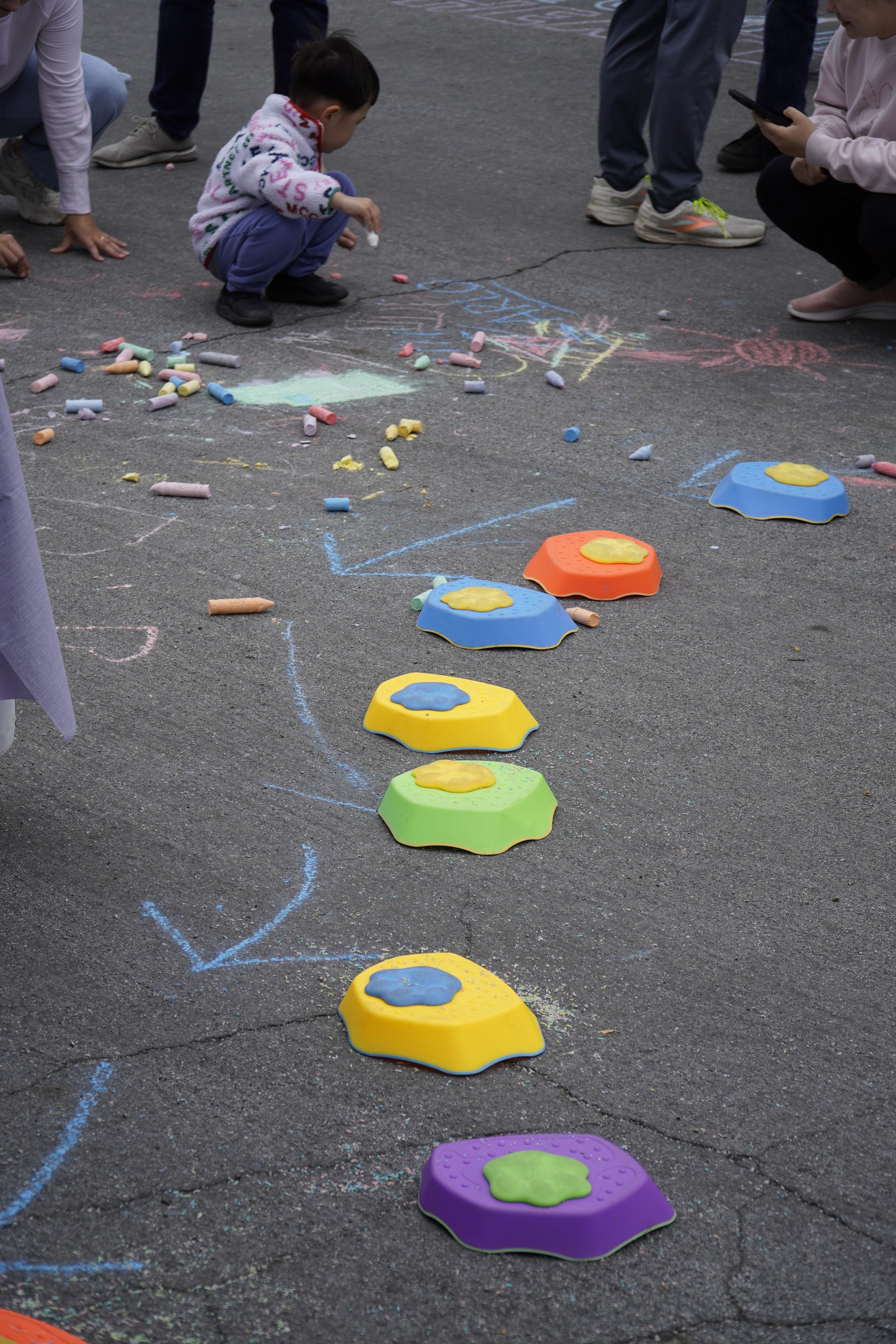 Children playing hopscotch on a paved ground with colorful stepping stones and chalk drawings, surrounded by several adults.