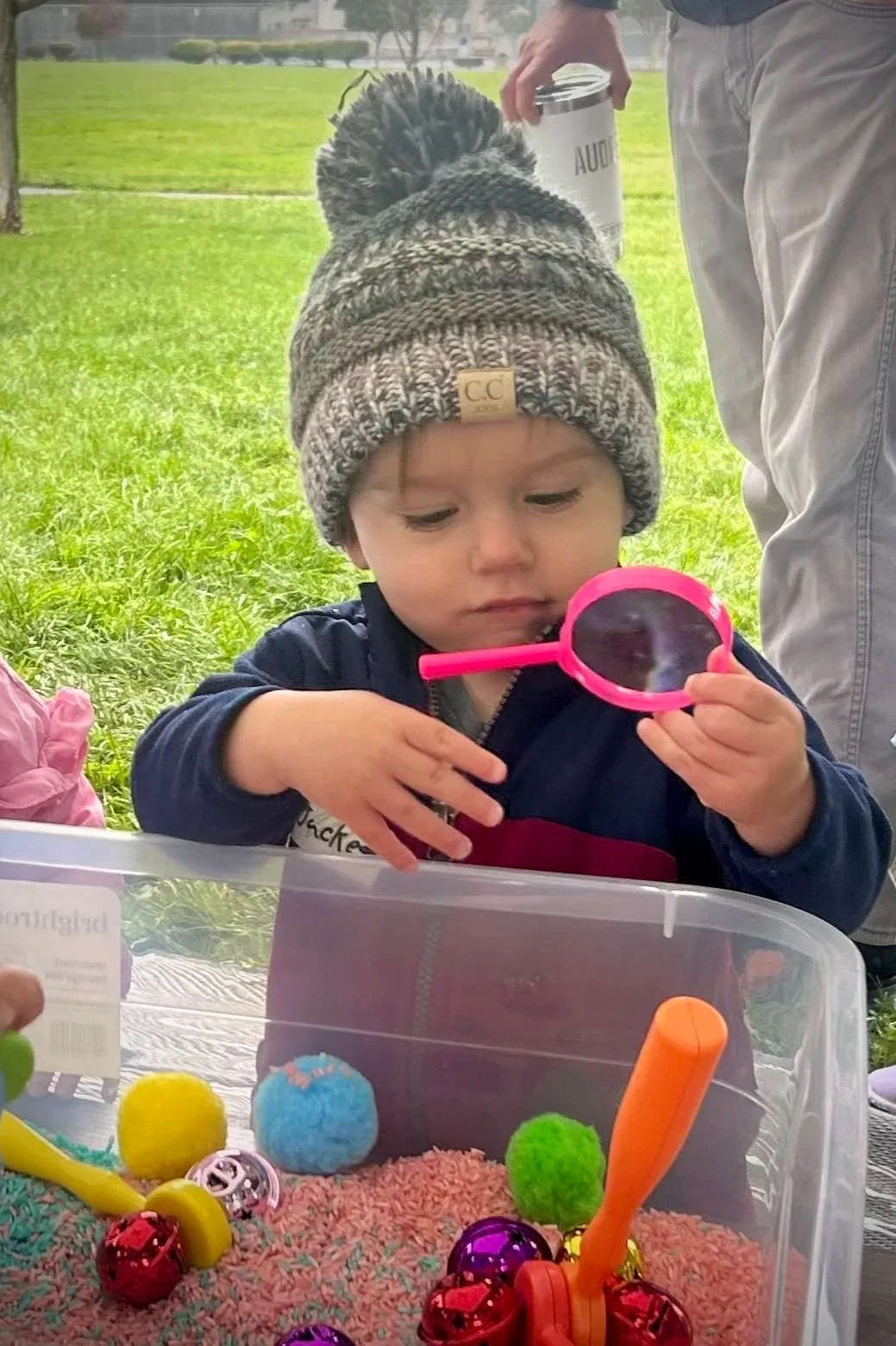 A young child wearing a gray knit beanie with a pom-pom and a navy jacket, holding a pink magnifying glass and inspecting colorful objects stored in a clear plastic container. The child is outdoors on a grassy area with a person in the background holding a white bucket.