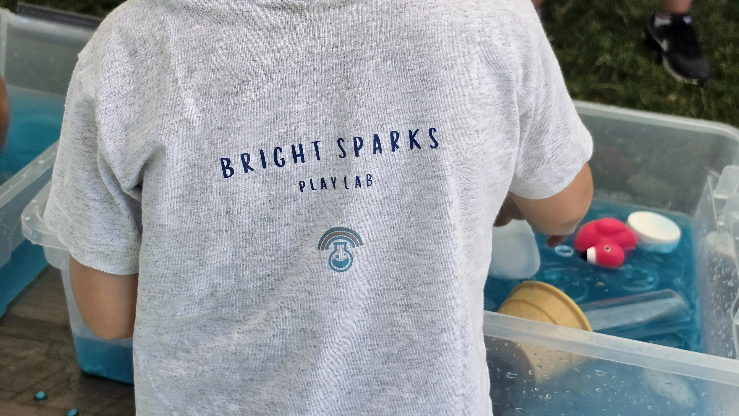 Child wearing a gray t-shirt with blue text that reads 'Bright Sparks Play Lab' and a small logo of a smiling face with a rainbow above it, standing near a sensory science treasure hunt with toys to pour, sort, measure and colorful objects, outdoors.