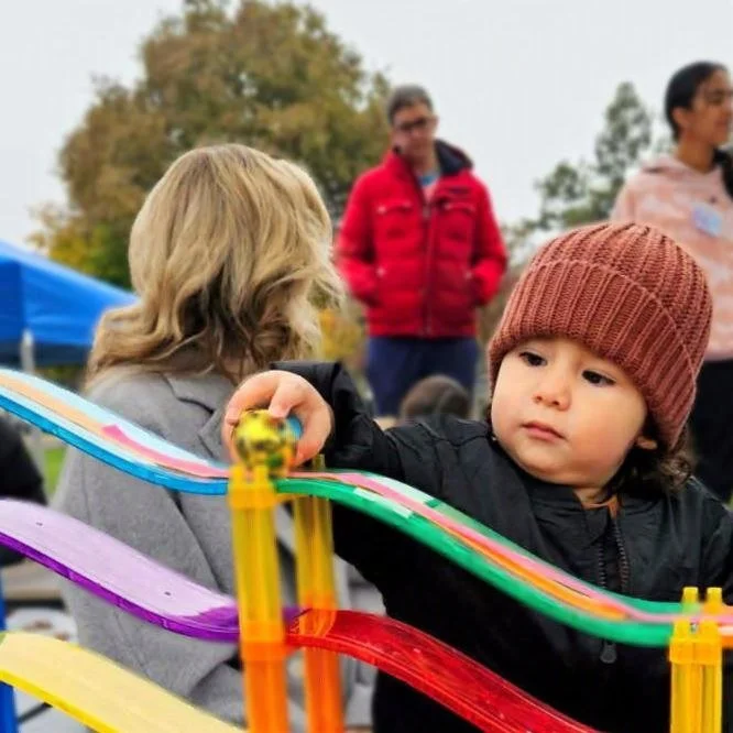 Young child in a brown beanie holding a jingle bell over a colorful magna-tile ramp, investigating the stick of the magnets to the bell. Other adults and children are in the background showing the community feel of the play lab.