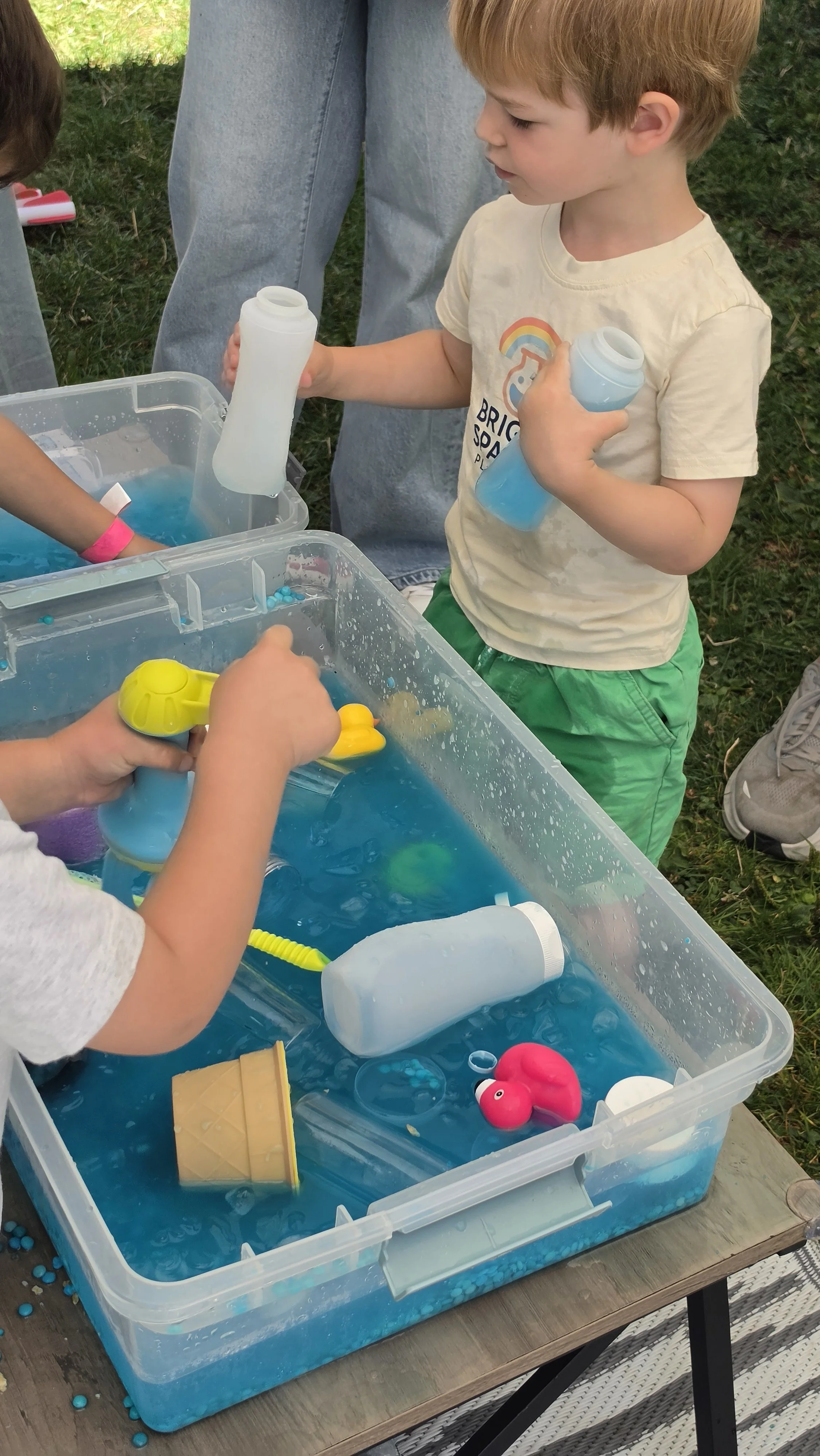 Children engaged in sensory science/STEM play in Mountain View. The children are experiencing ice as it melts while rescuing frozen rubber ducks, flamingos, and collecting treasure. They are holding bottles of warm water to melt the ice.