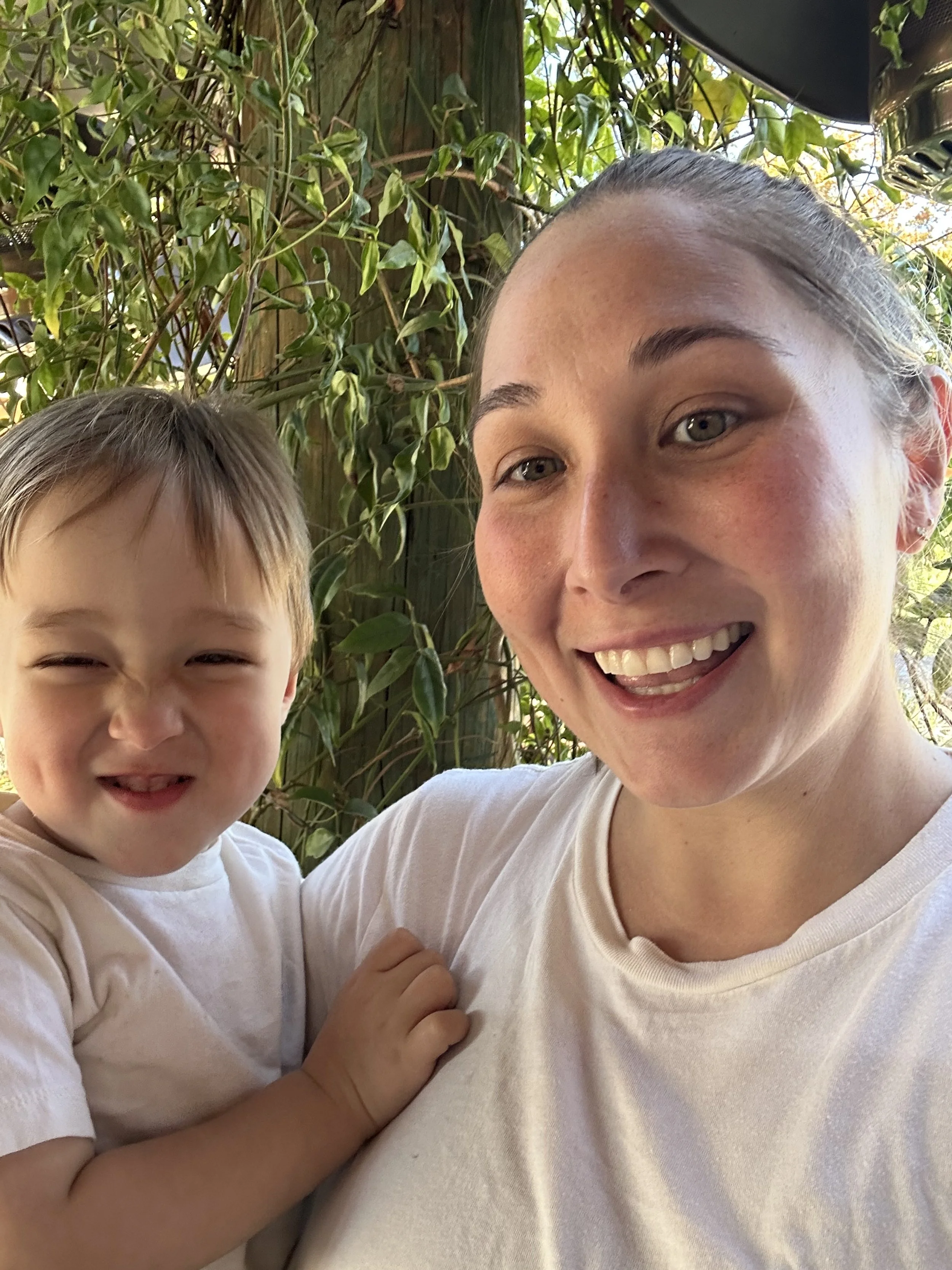 A woman with light skin, light brown hair, and blue eyes smiling while holding a young boy with light skin, light brown hair, and brown eyes. They are outdoors with green foliage in the background.