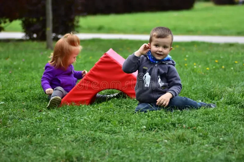 two-toddlers-boy-gray-hoodie-girl-purple-jacket-sitting-grass-red-foam-playground-block-enjoying-outdoor-play-404815694.webp