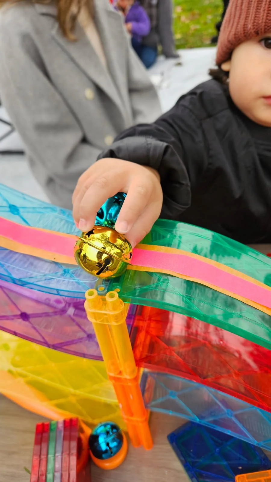 Child playing with colorful translucent marble run toy outdoors, with a gold-colored bell on top.