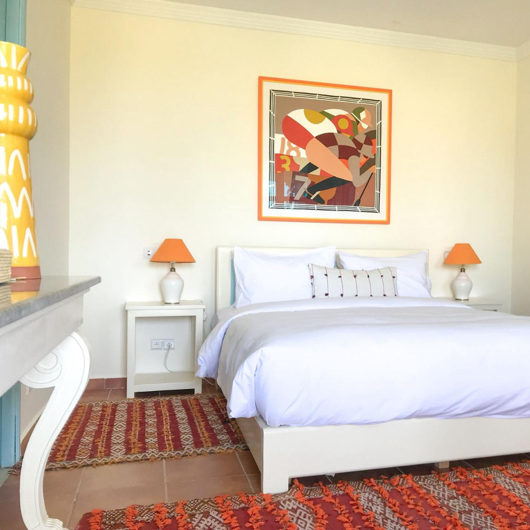 A bedroom with a white bed, orange lamps on white nightstands, a colorful abstract art piece above the bed, and patterned rugs on the floor.