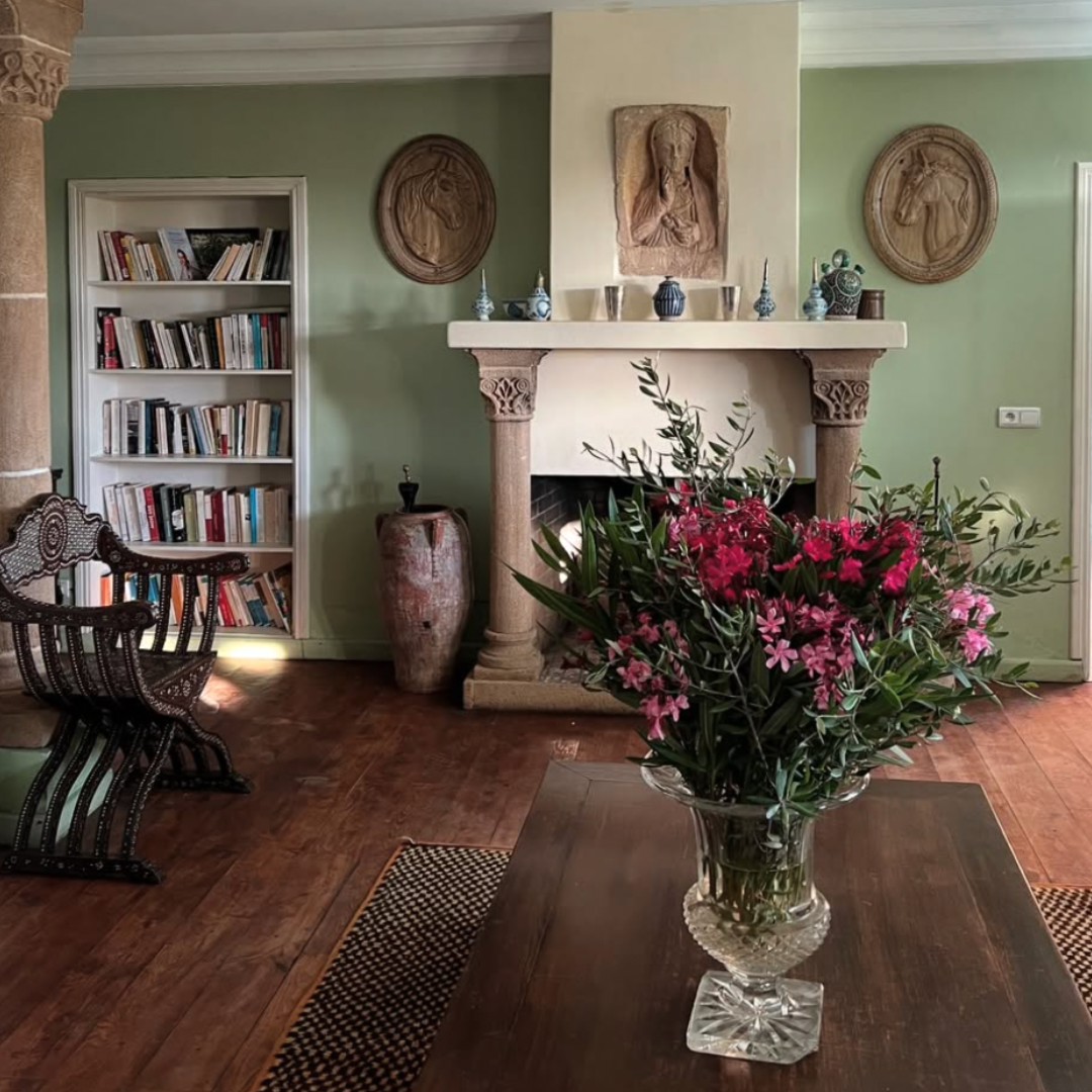 A cozy living room featuring a stone fireplace with decorative columns, framed masonry art on the wall, a built-in bookshelf filled with books to the left of the fireplace, a large vase with a plant to the right, a wooden table with a glass vase fill