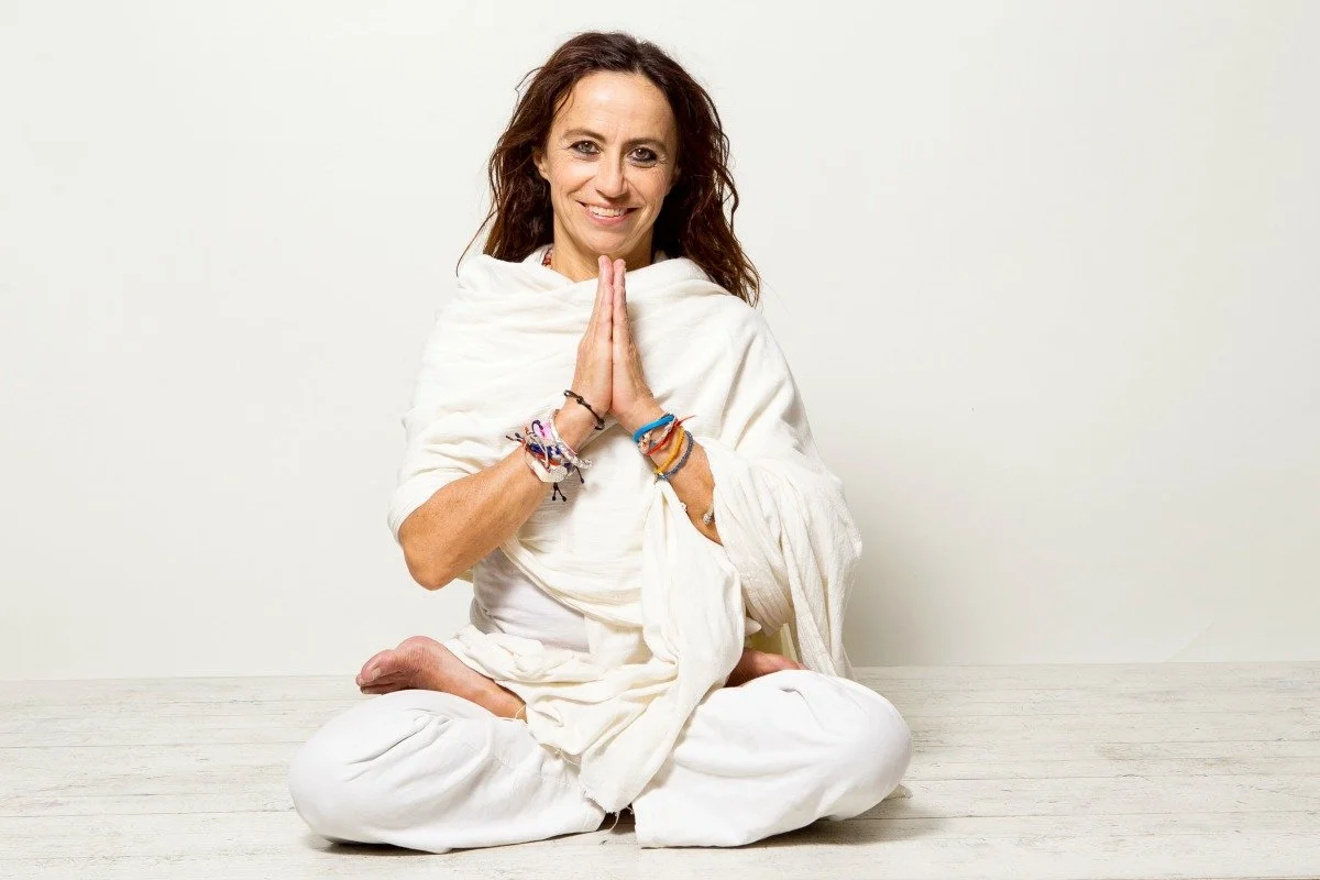 A woman sitting cross-legged on the floor with her hands in a prayer position, smiling, against a plain white background.