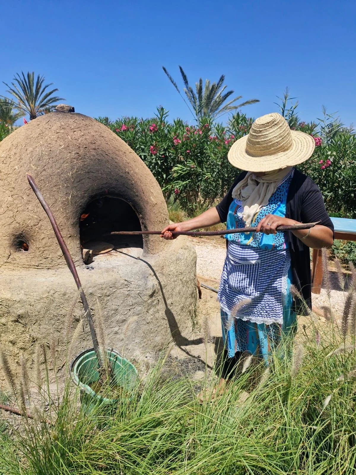 A person wearing a wide-brimmed straw hat and colorful clothing is using a long stick to tend to a clay oven outdoors, surrounded by greenery and pink flowers under a clear blue sky.