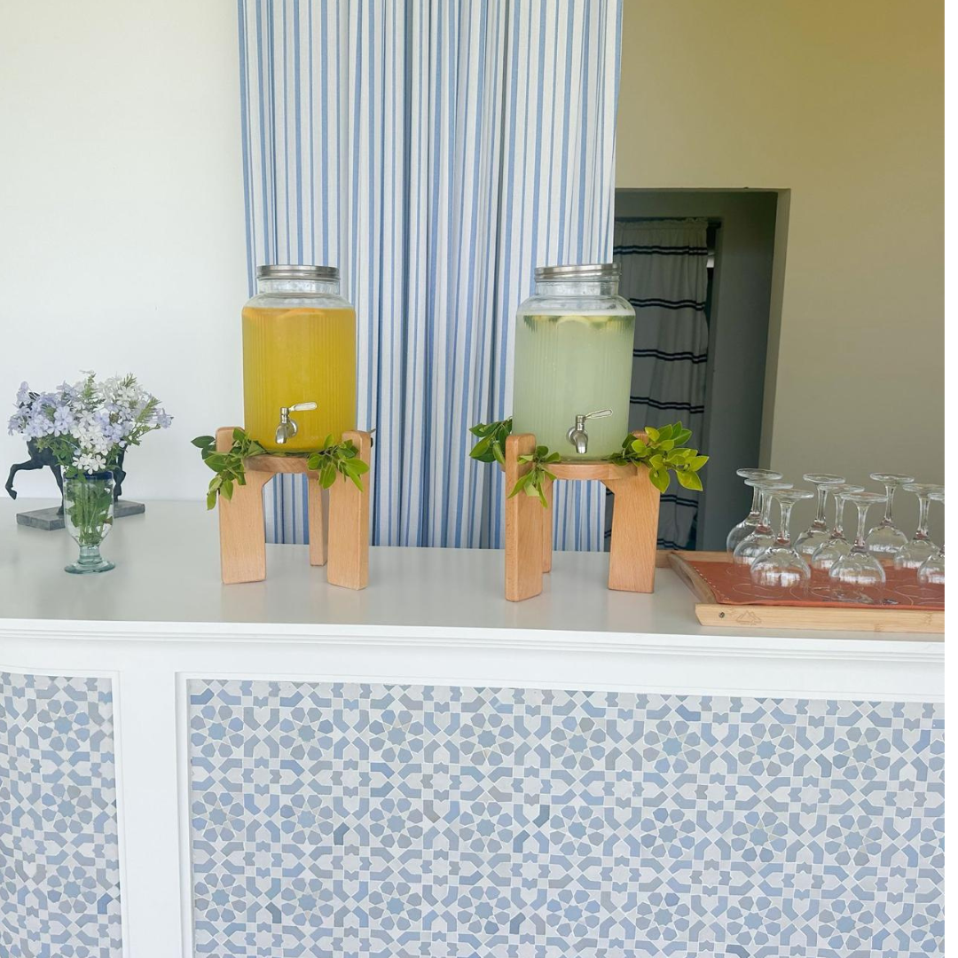 Two large glass beverage dispensers with spigots, one filled with yellow drink and the other with light-colored drink, on wooden stands decorated with green leaves, set on a white counter with a patterned front. A tray with upside-down glasses is on 