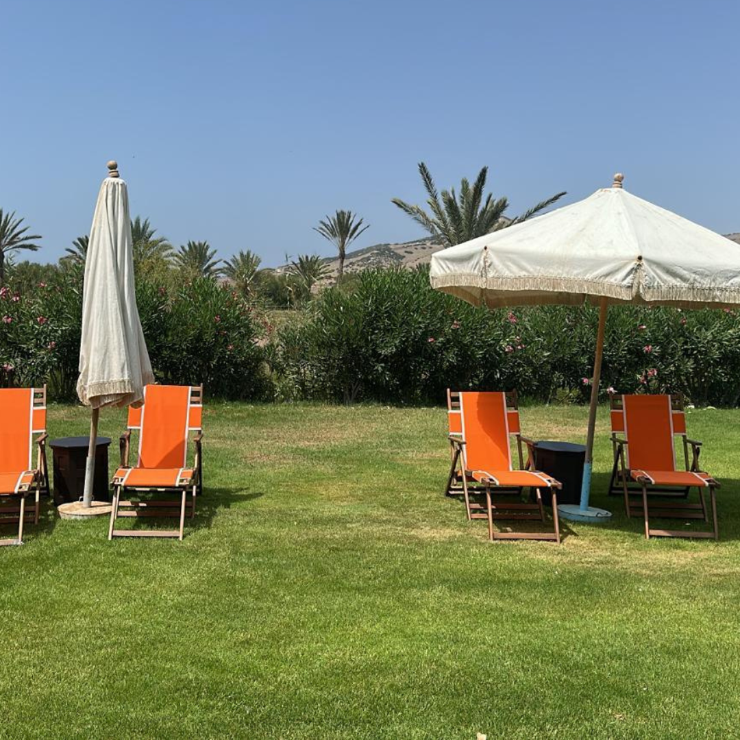 Patio scene with four orange lounge chairs with matching small side tables, two umbrellas (one closed and one open), lush green grass, bushes with pink flowers, and palm trees in the background under a clear blue sky.