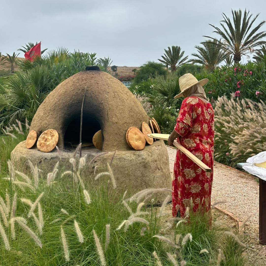 A woman in a floral red dress and wide-brimmed straw hat is baking bread in a traditional clay oven outdoors, surrounded by lush green plants and palm trees.