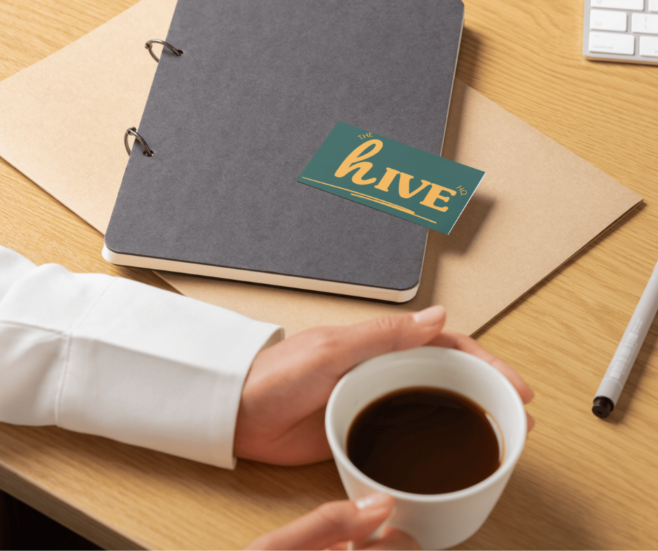 A person holding a cup of black coffee at a wooden desk with a notebook, a pen, and a keyboard.