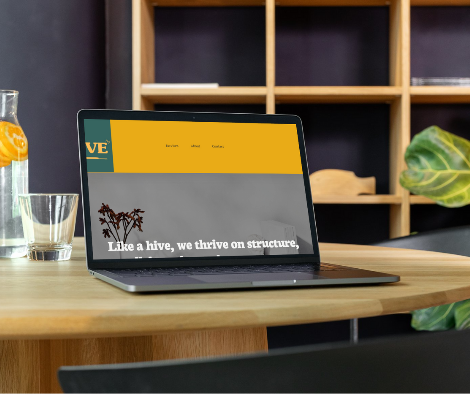 A laptop on a wooden table displaying a website with a yellow header and the text 'Like a hive, we thrive on structure.' In the background, there is a wooden shelf, and on the table, there is a glass pitcher with lemon slices and an empty glass. A green plant leaf is visible on the right side of the image.