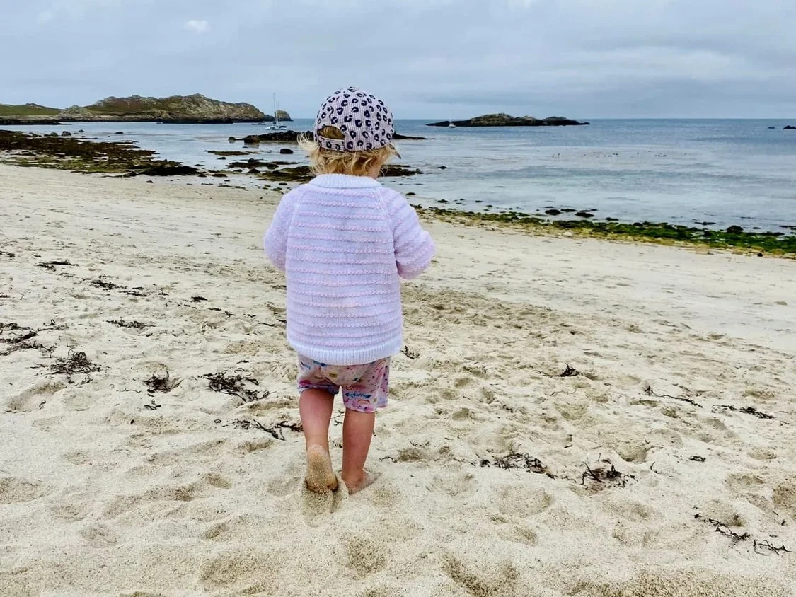 A young child with blonde hair, wearing a patterned hat, light pink sweater, and colorful shorts, walking on a sandy beach toward the water.