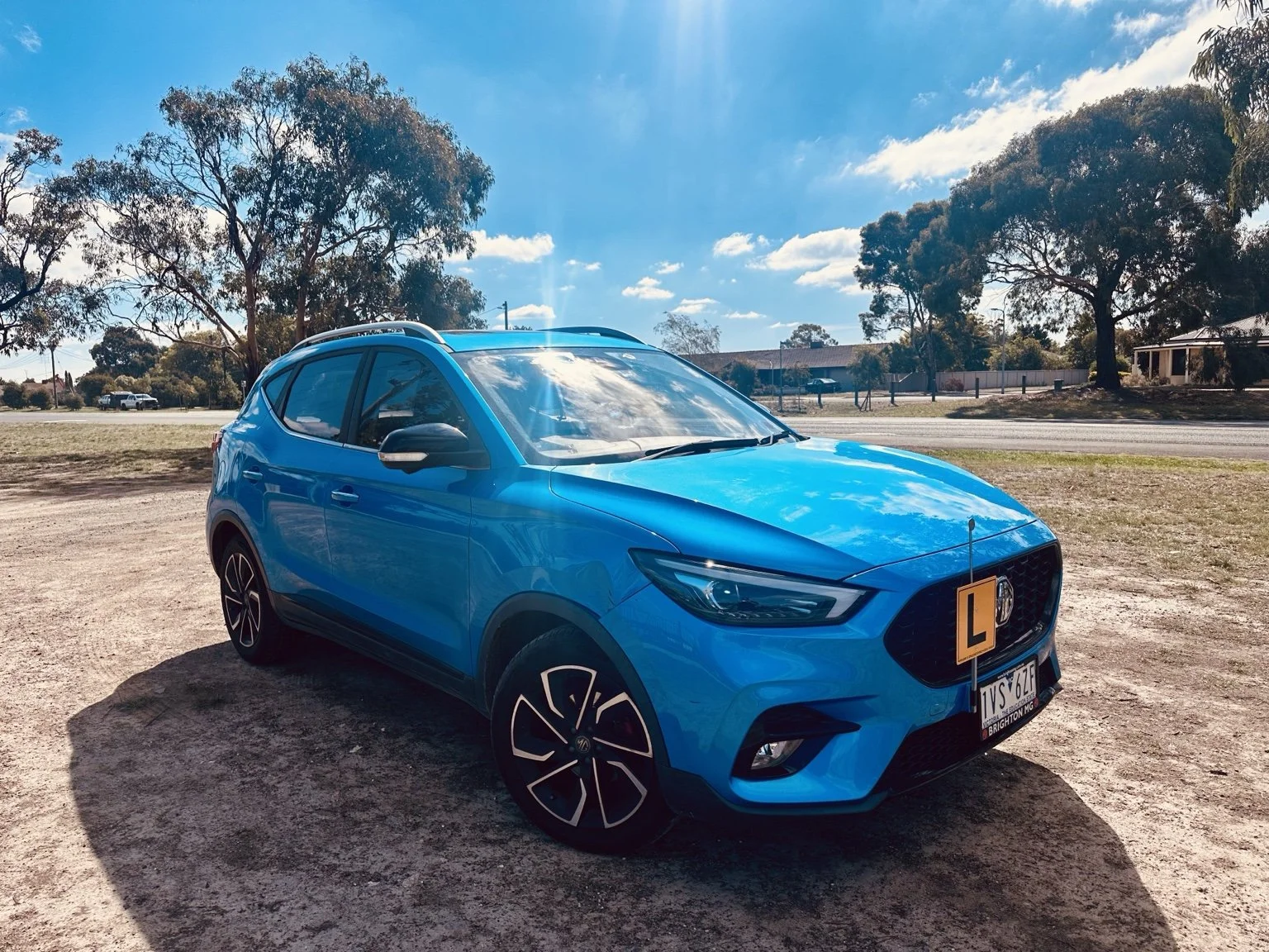 A bright blue car with an 'L' learner driver plate parked on a dirt area under a clear sky with some clouds, with trees and houses in the background.