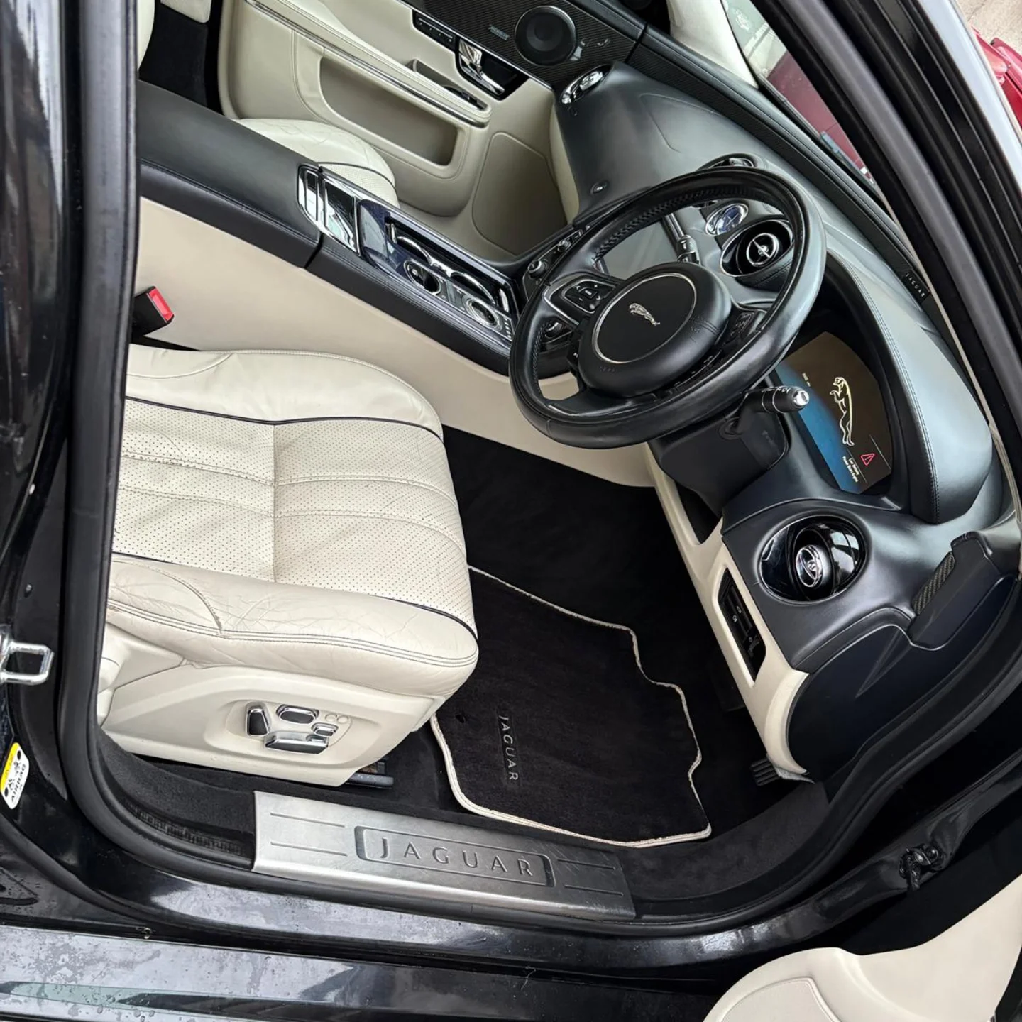 Interior of a Jaguar vehicle with cream leather seats, black and cream dashboard, steering wheel with Jaguar logo, digital display, and black floor mats labeled 'Jaguar'.