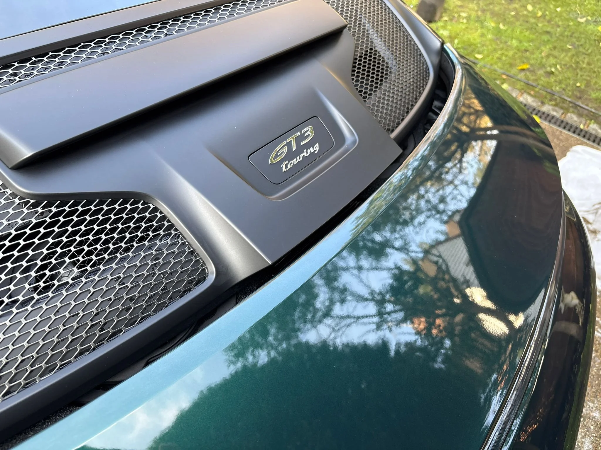 Close-up of a car's hood showing a 'GT3 Touring' badge and a large front grille with honeycomb pattern, with reflections of trees and sky on the shiny green surface.