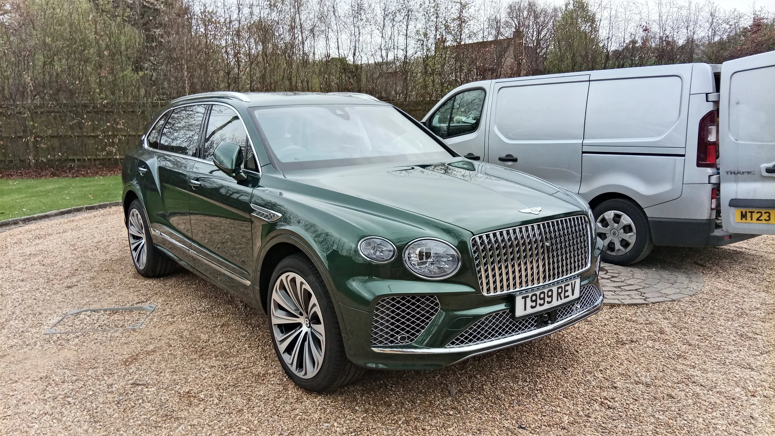 A dark green luxury station wagon and a silver cargo van parked on a gravel driveway, with a background of trees and a wooden fence.