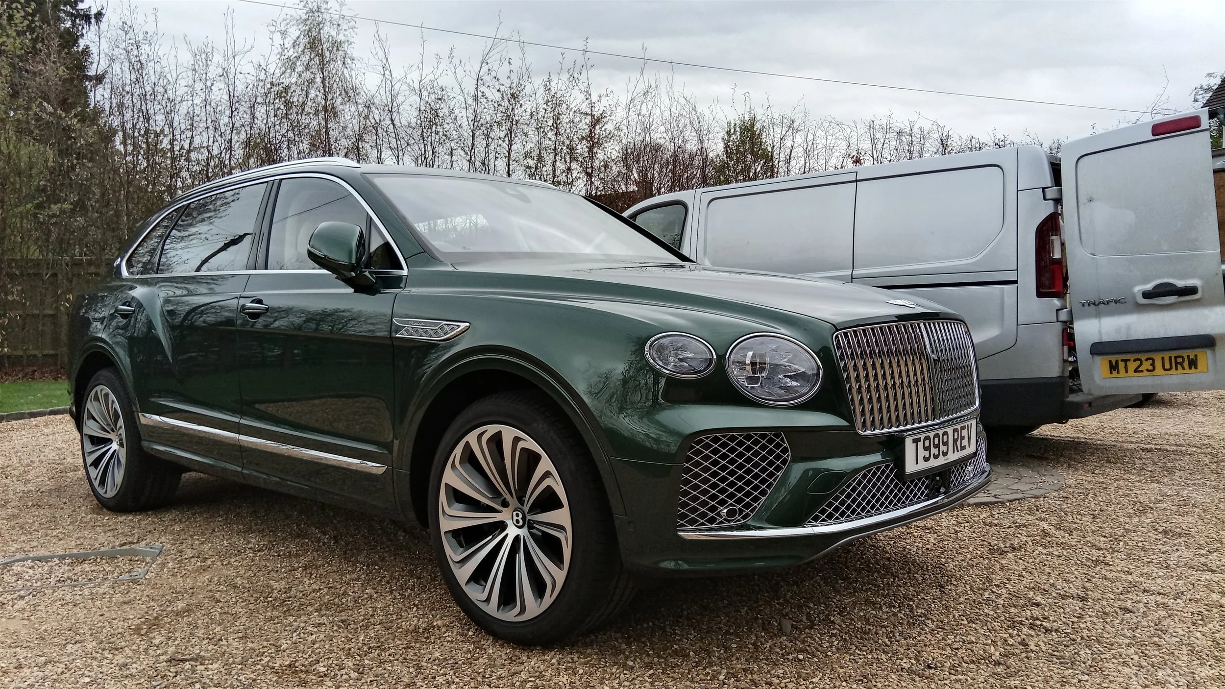 A green luxury SUV Bentley parked next to a white van on a gravel surface with leafless trees and overcast sky in the background.
