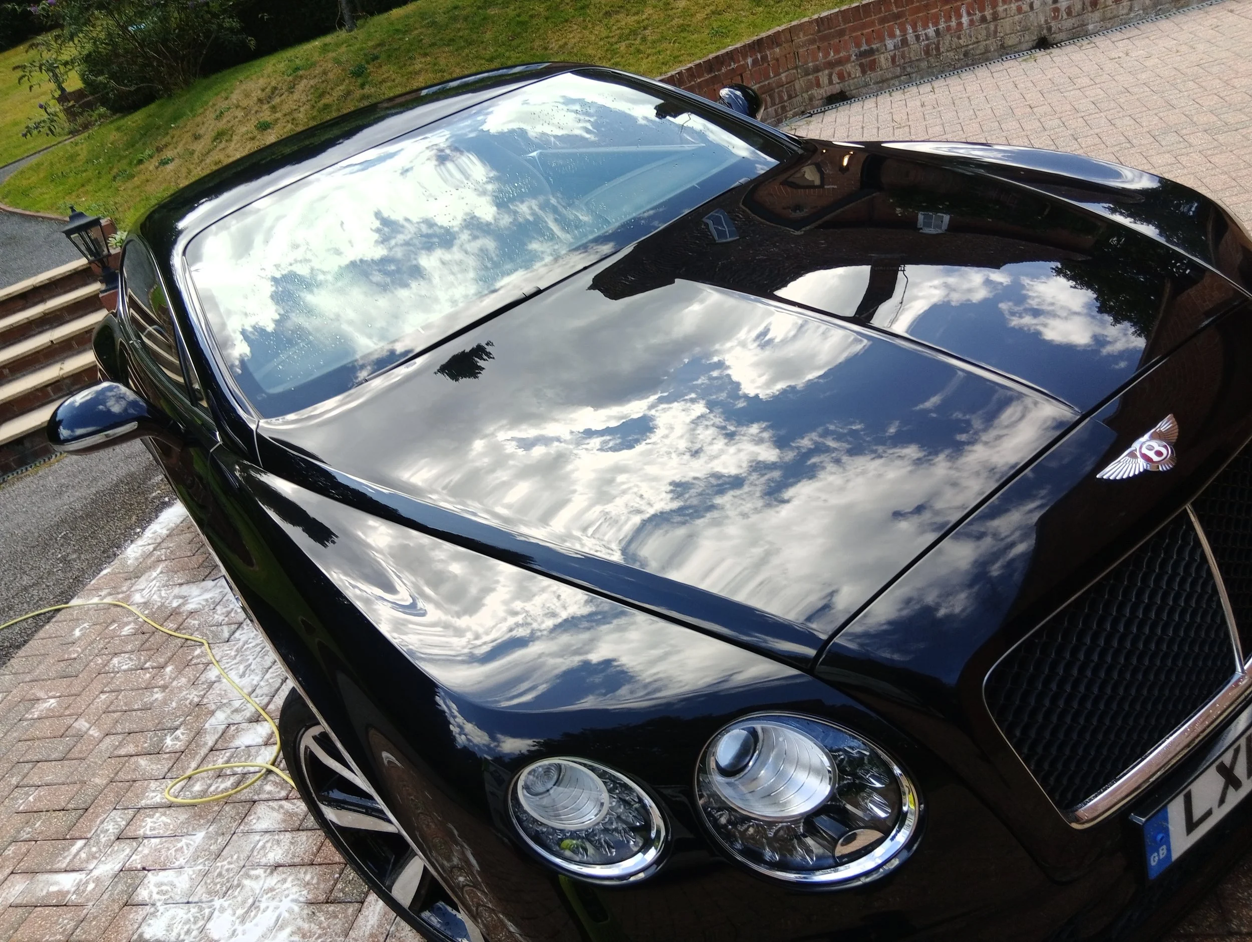 Black Bentley car parked on a brick driveway, reflecting the cloudy sky and clouds on its hood and windshield.