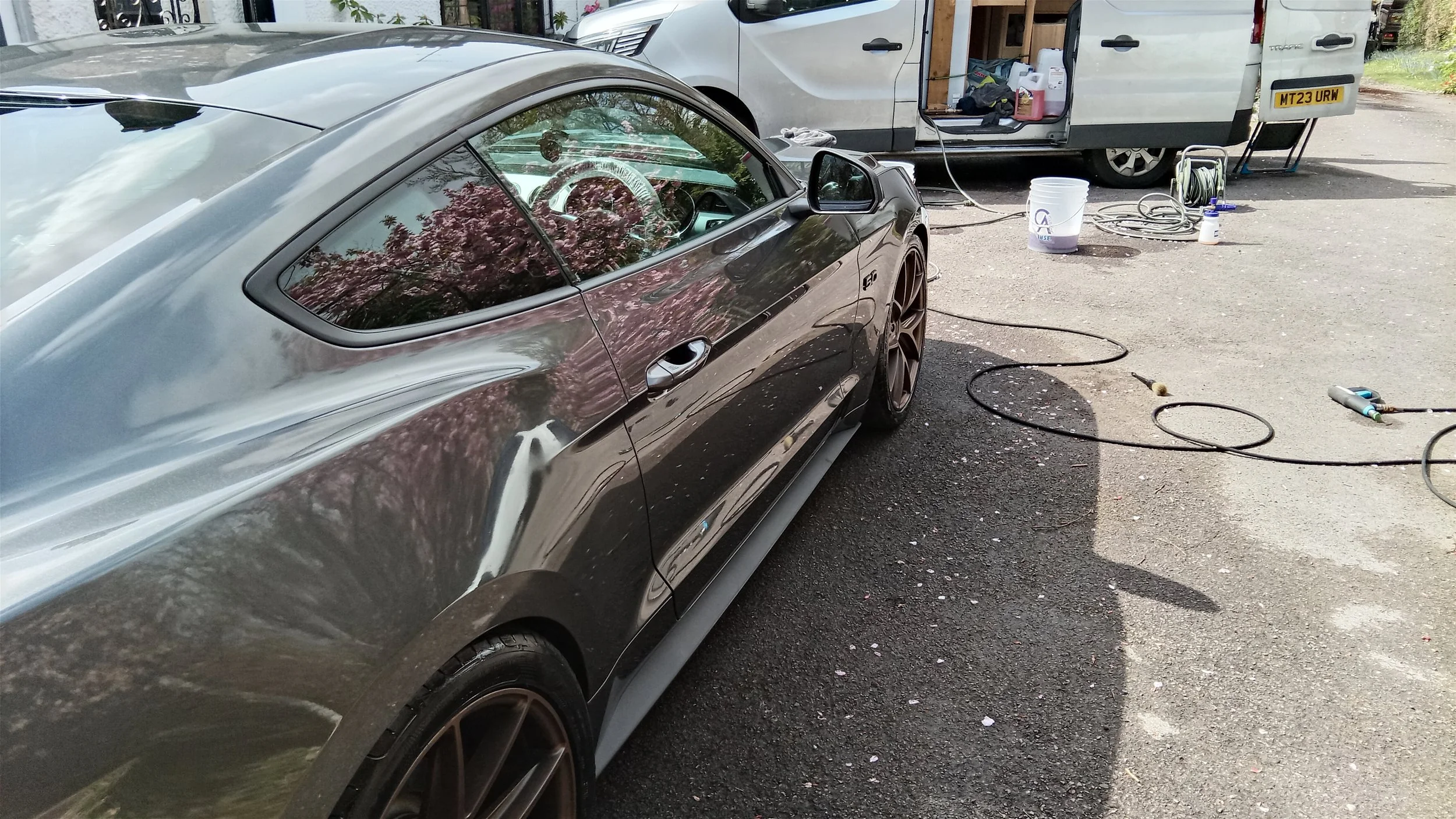 A black sports car is being cleaned or polished outdoors near a white van with an open side door. The scene includes cleaning equipment such as hoses, buckets, and brushes on the ground.