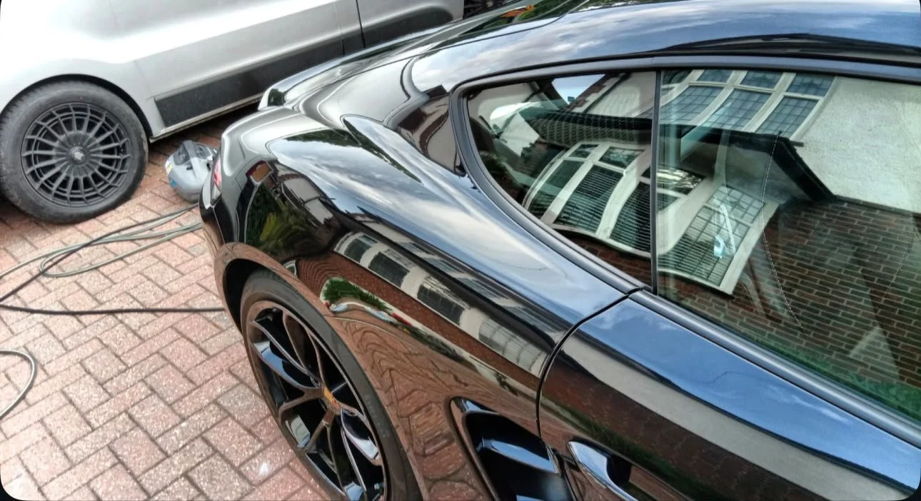 Black sports car parked on a brick driveway, reflecting a house with white windows on its surface, with a gray car nearby and an electric blower on the ground.