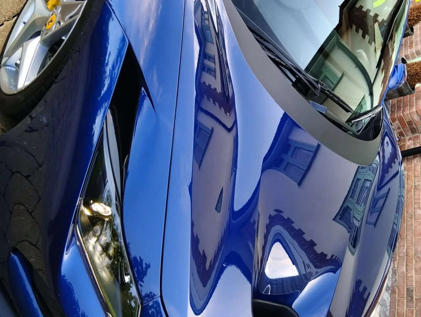 A close-up of a shiny blue sports car parked next to other vehicles, with a brick wall and residential buildings reflected on its surface.