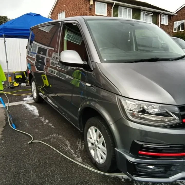 A gray van parked on a wet driveway with soap and water, being washed under a blue canopy tent, with a house in the background.