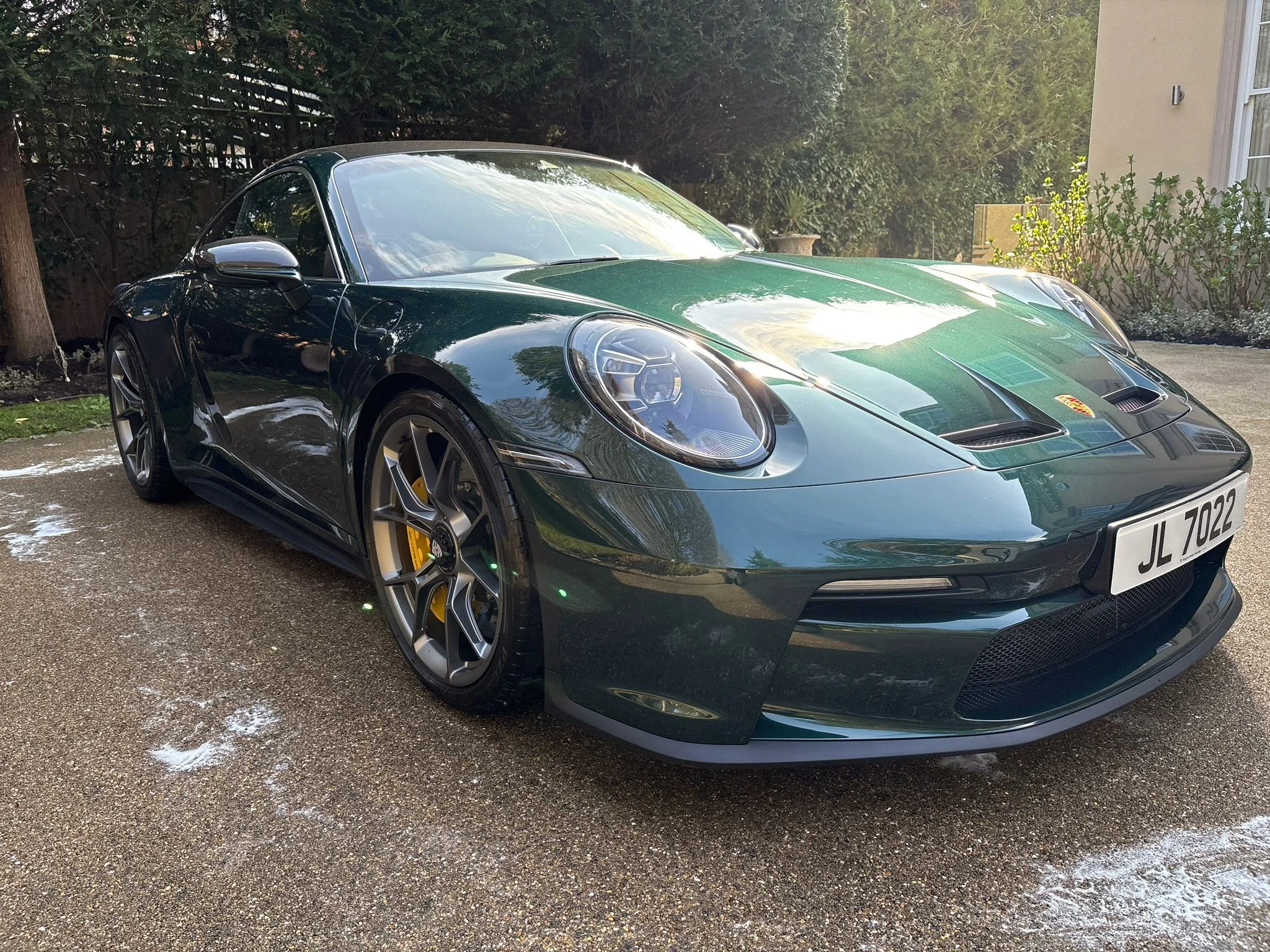 A dark green Porsche sports car parked on a driveway with trees and a house in the background.