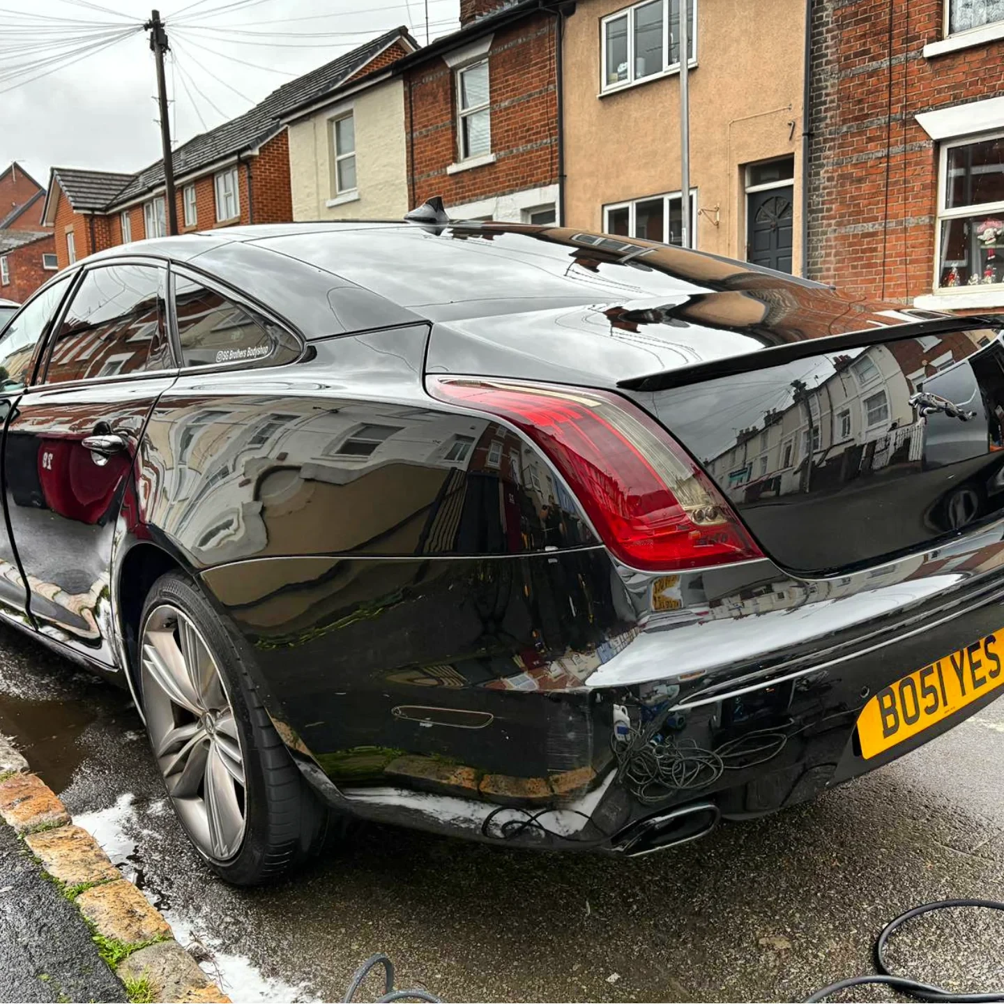 A black luxury sedan parked on the street with a reflection of a row of houses on its surface. The car has a yellow license plate that reads 'BOSIYES' and is connected to a small black electrical device near the rear wheel. The street has a wet surfa