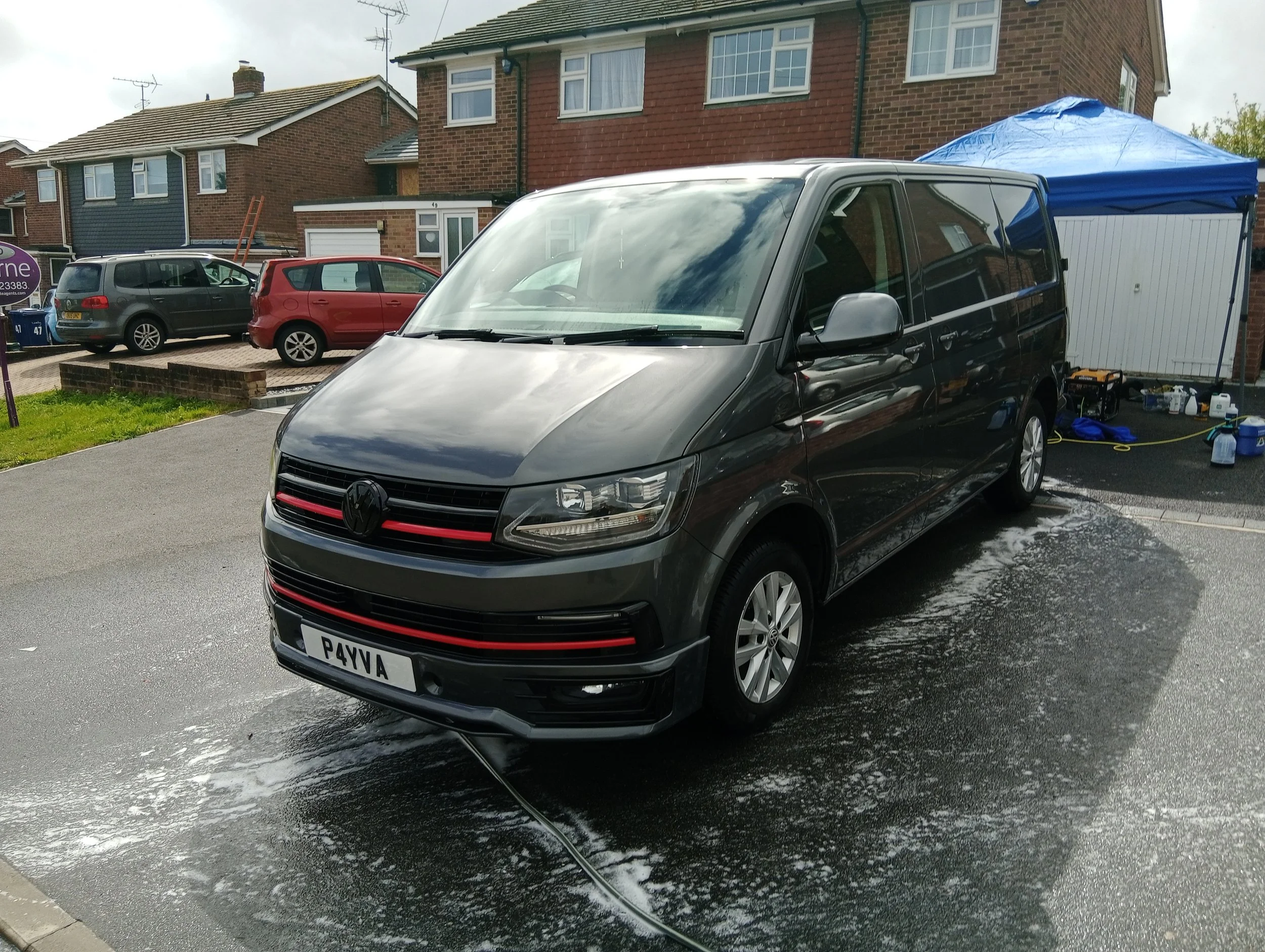 A black Volkswagen van parked on a driveway with soap suds and water, indicating it is being washed. In the background, there are several other parked cars, a brick house, and a blue pop-up tent with cleaning supplies underneath.